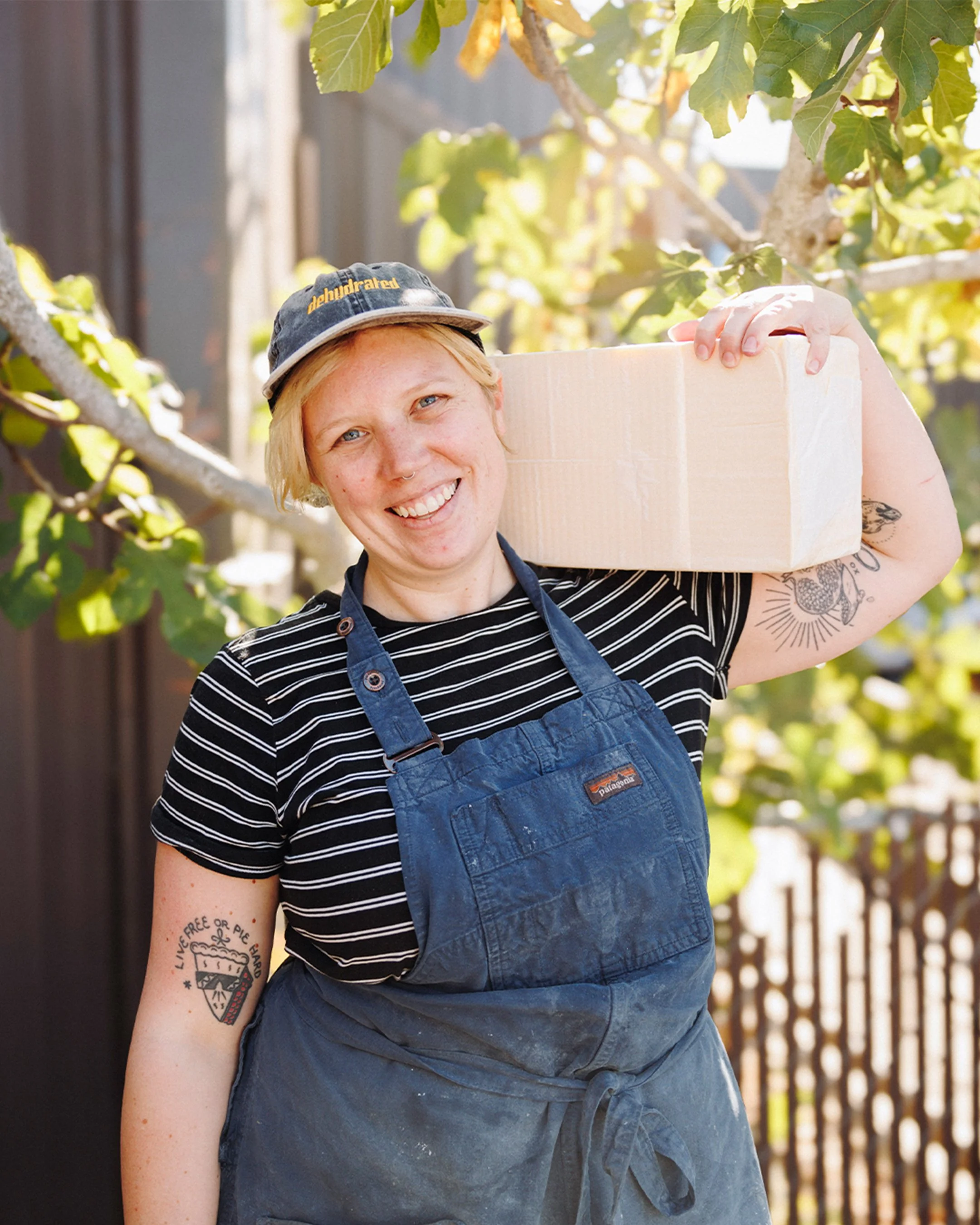 A smiling woman with short blonde hair and tattoos on her arms, wearing a black and white striped shirt, a blue apron, and a hat, holding a cardboard box on her shoulder outdoors with trees and sunlight in the background.