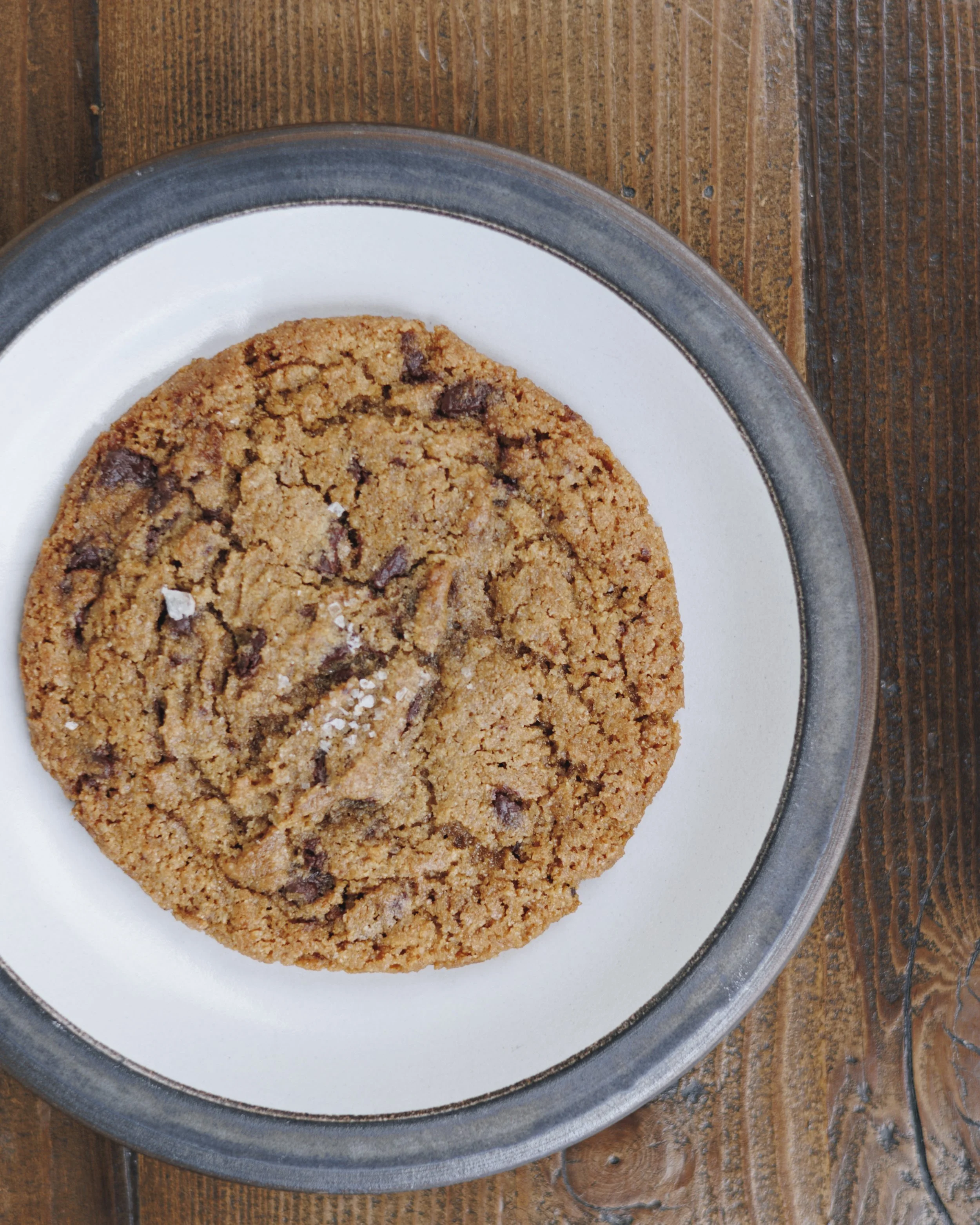 A chocolate chip cookie on a white plate with a gray rim, placed on a wooden table.