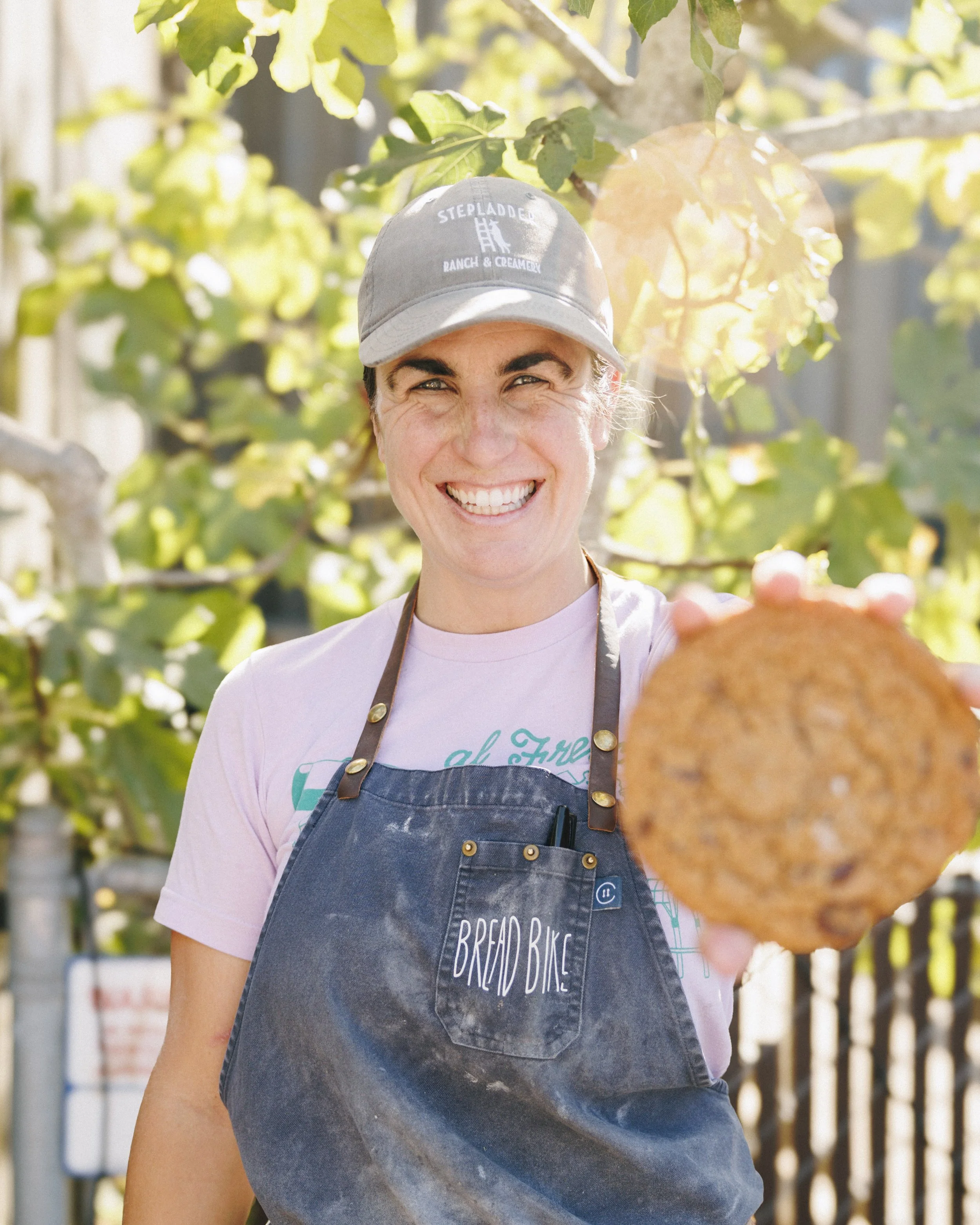 A woman smiling and holding out a cookie towards the camera, wearing a gray baseball cap, a pink t-shirt, and a blue apron with 'Bread Bike' embroidered on it. She is outdoors with trees in the background.