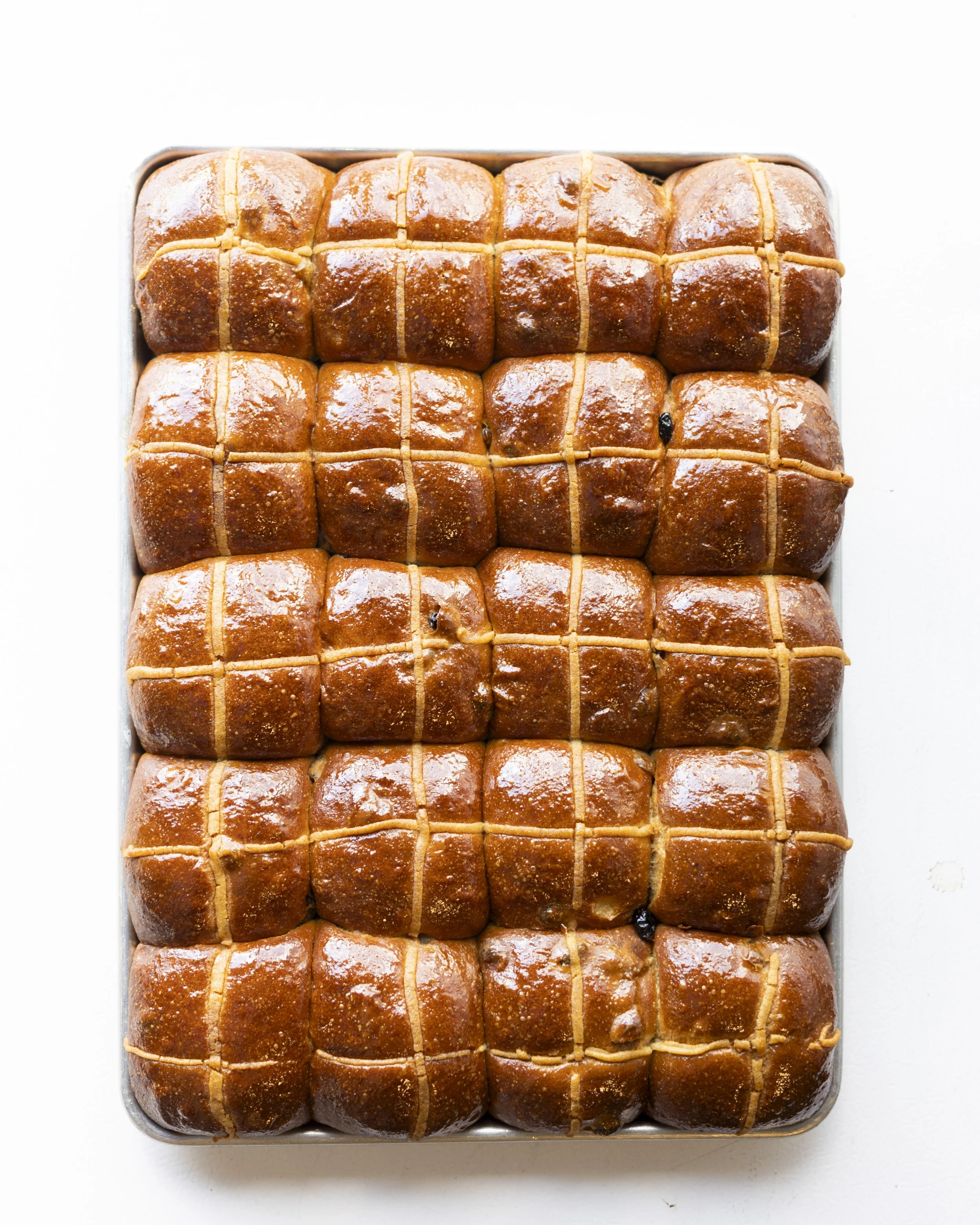 A tray of glazed hot cross buns arranged in four rows of four, each with a cross on top.