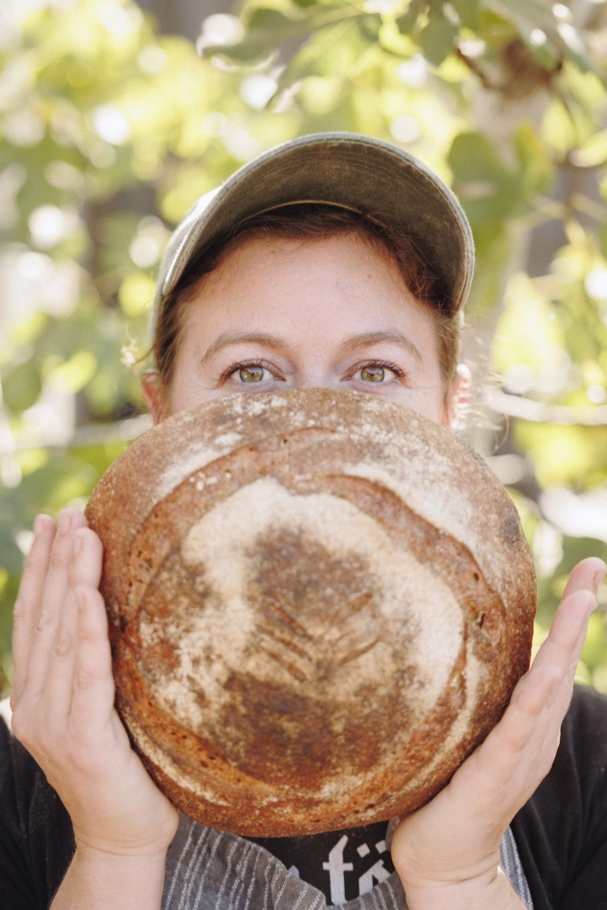 Person holding a loaf of bread in front of their face, showing only their eyes and part of their hat. The background is outdoor with green leaves.