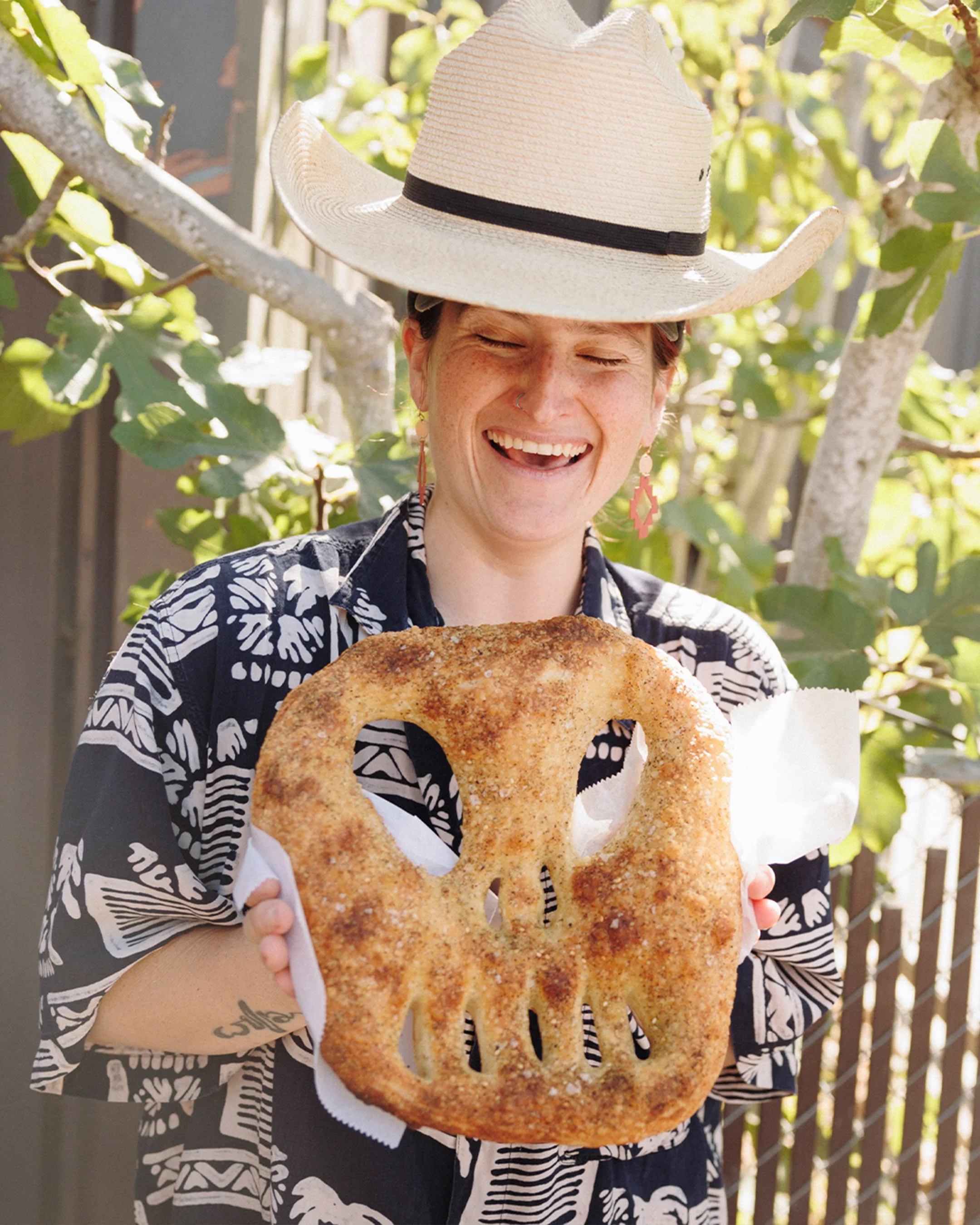 A woman wearing a large straw hat and a patterned shirt smiling while holding a large pretzel shaped like a skull outdoors.