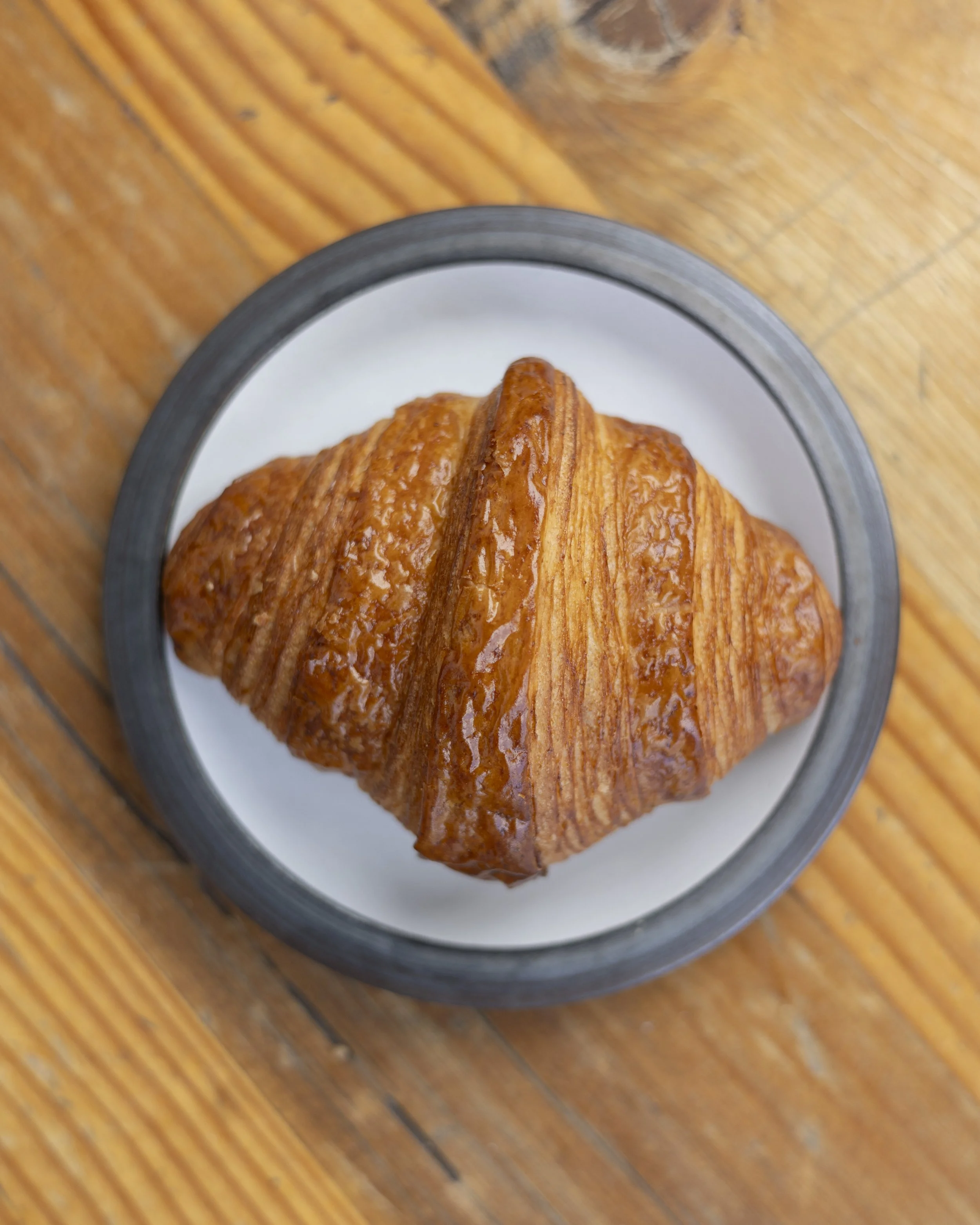 A flaky, golden-brown croissant on a white plate with a black rim, placed on a wooden surface.