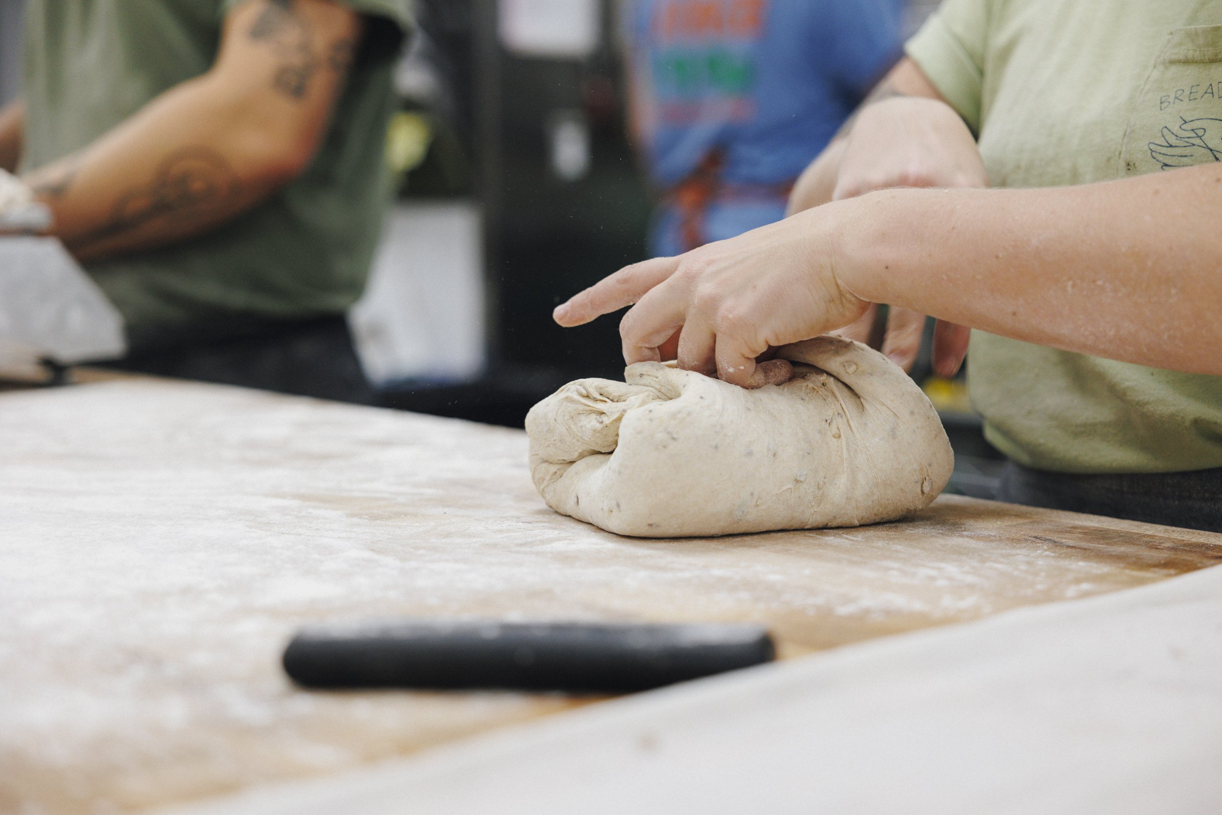 Person kneading dough on a floured wooden surface in a bakery or kitchen.
