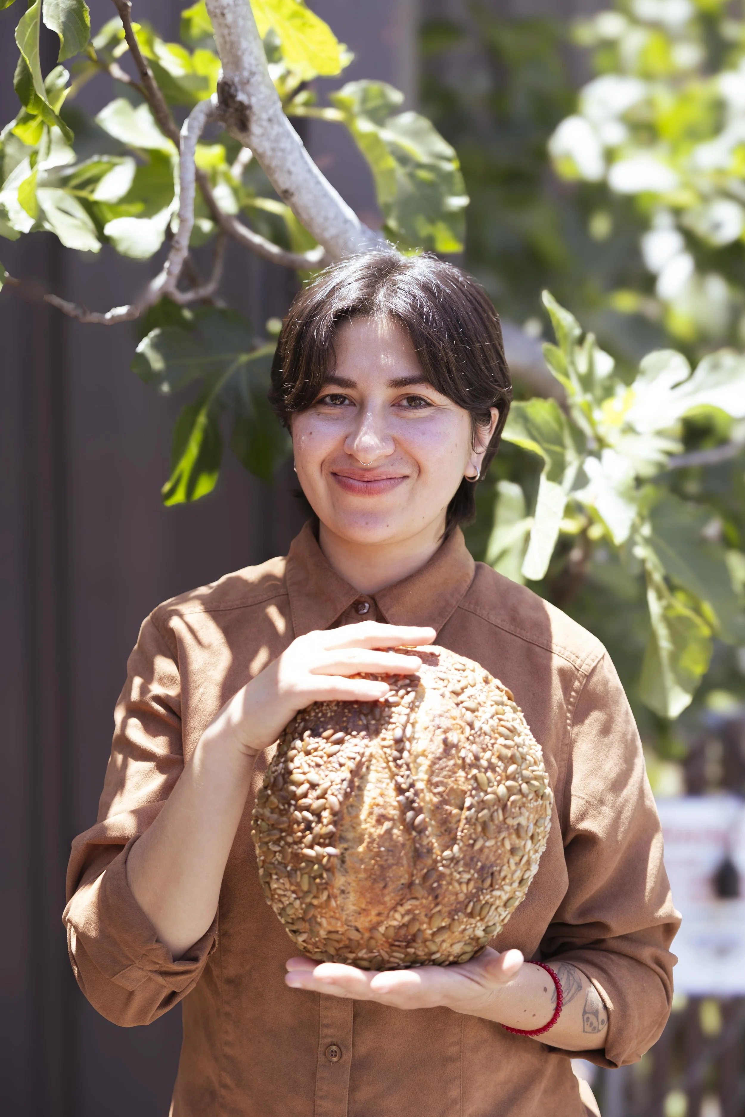 A woman with short dark hair smiling while holding a large round bread covered in seeds outdoors.