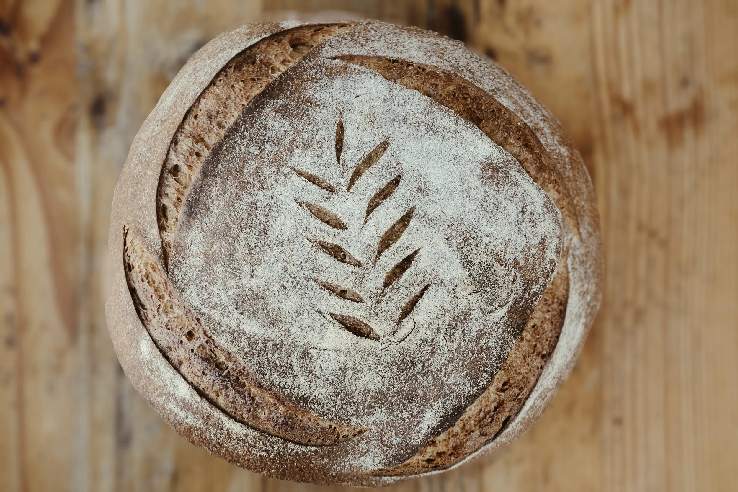 Close-up of a round loaf of bread with a leaf-shaped cutout on the top, dusted with flour, on a wooden surface.