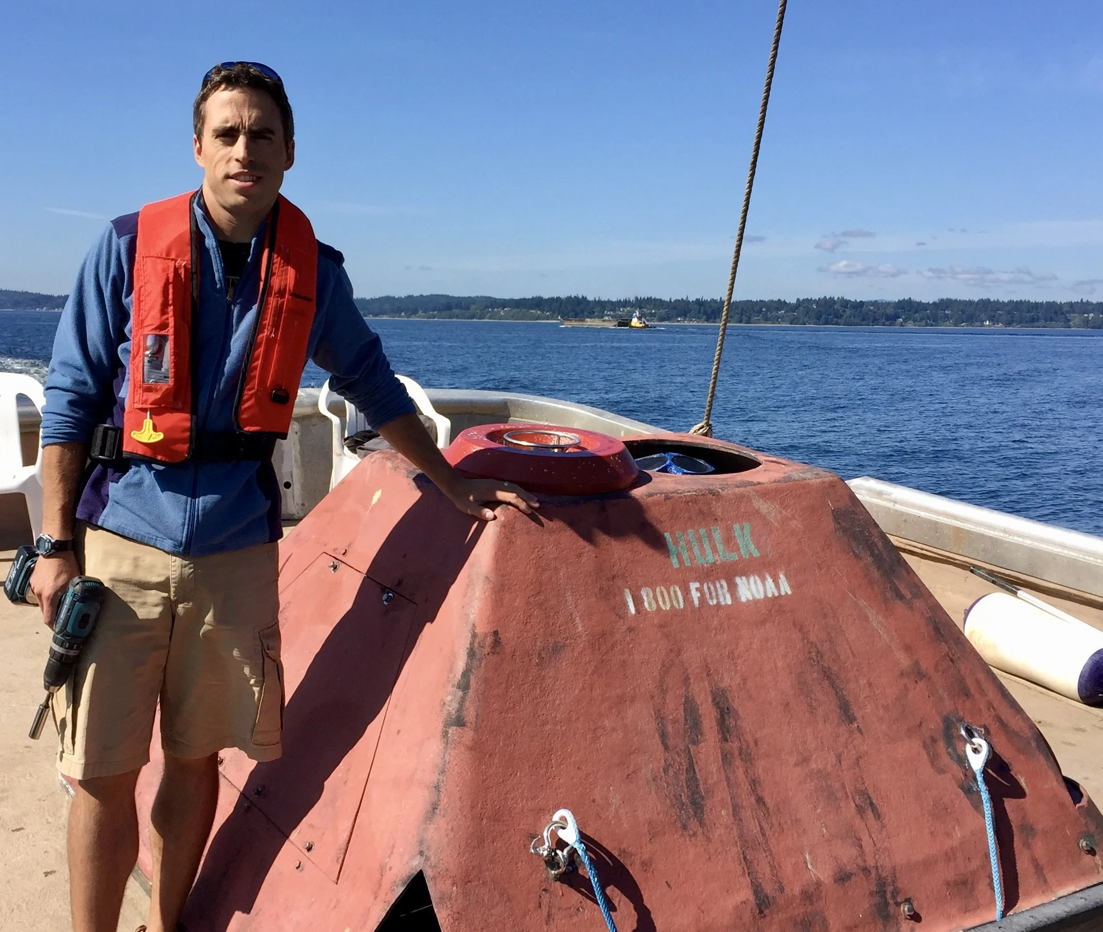 A man in a blue jacket and beige shorts standing on a boat near a red submarine with the name "HULK" painted on it, holding a power drill, with water and distant land in the background.