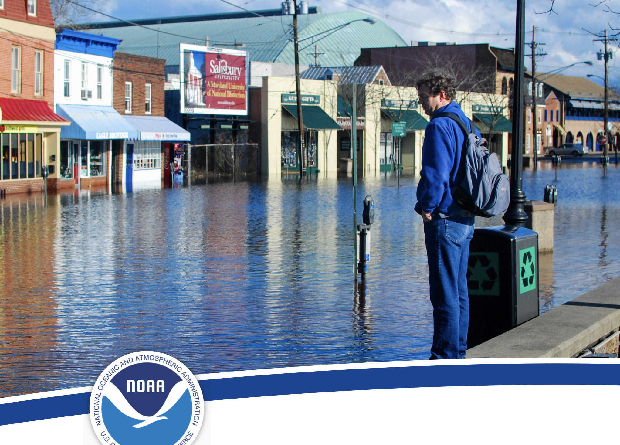 A person wearing a blue hoodie and jeans standing on a flooded sidewalk next to a trash can with a recycling symbol, looking at floodwaters caused by severe flooding in an urban area with buildings and shops in the background.