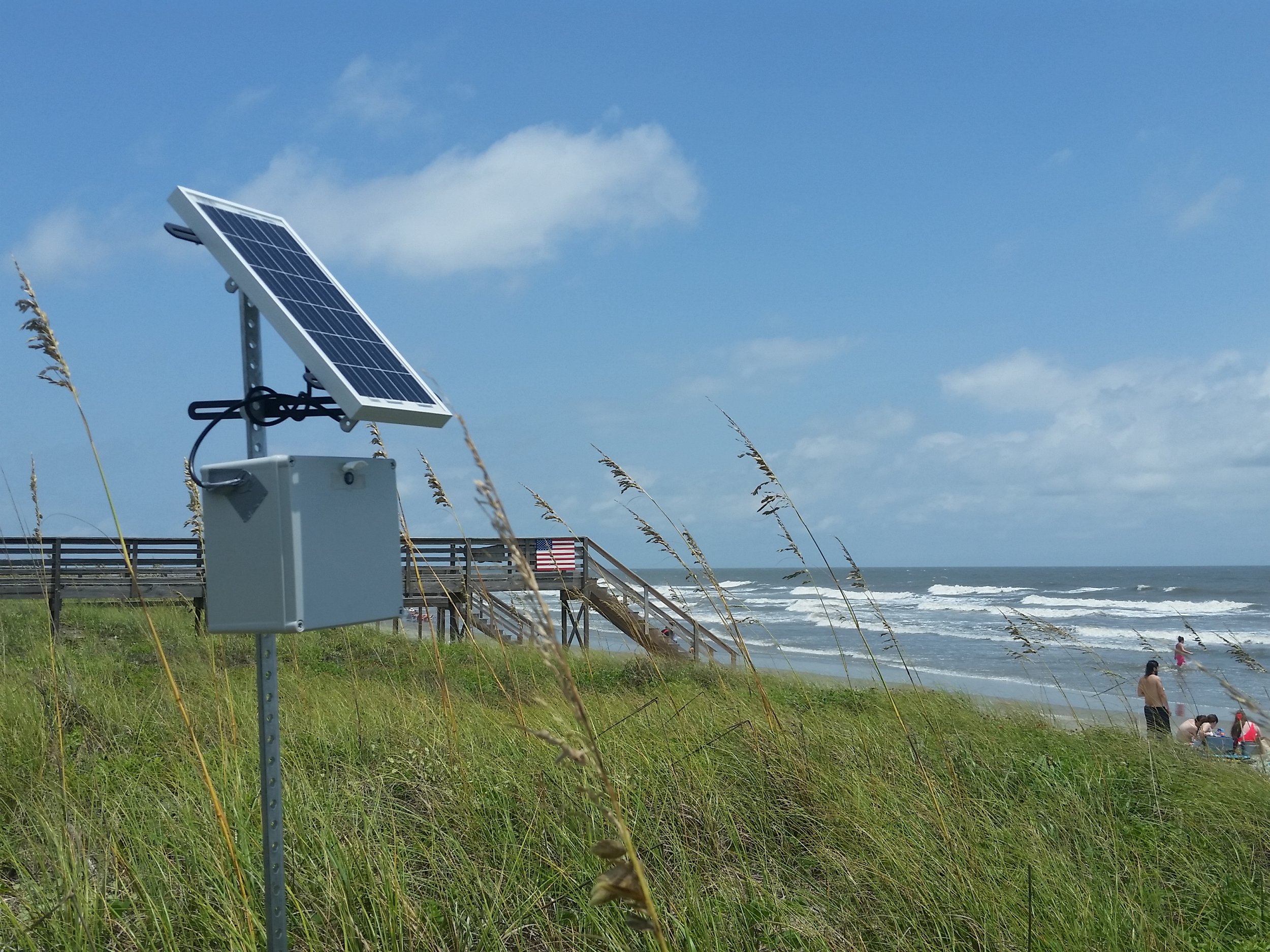 A camera and solar panel installed on a pole near a grassy area overlooking a beach with people in the water and a wooden staircase leading down to the shore, under a partly cloudy sky.