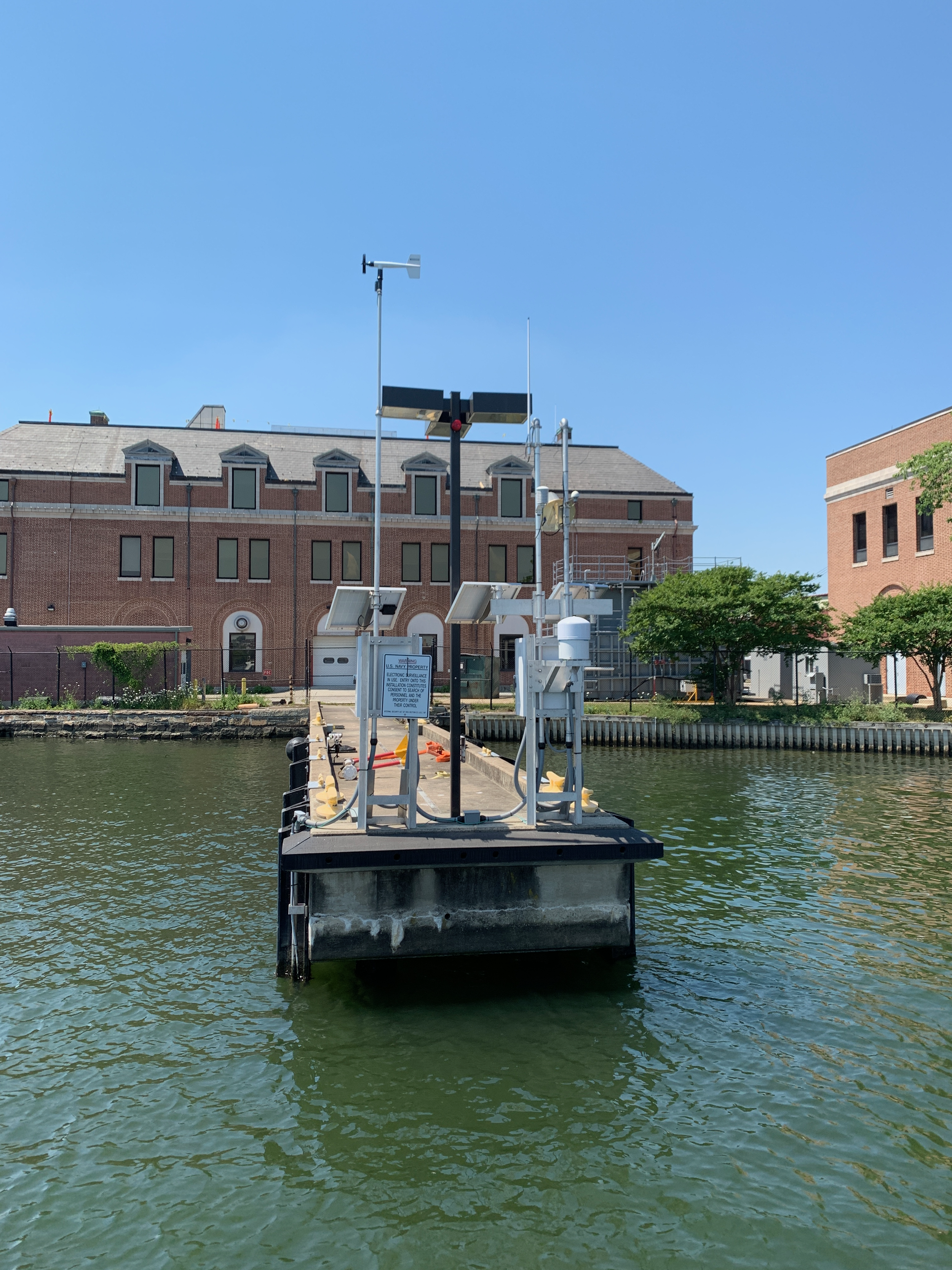 A NOAA water level station equipped with various sensors, antennas, and solar panels, situated on a body of water, with a brick building and trees in the background under a clear blue sky.