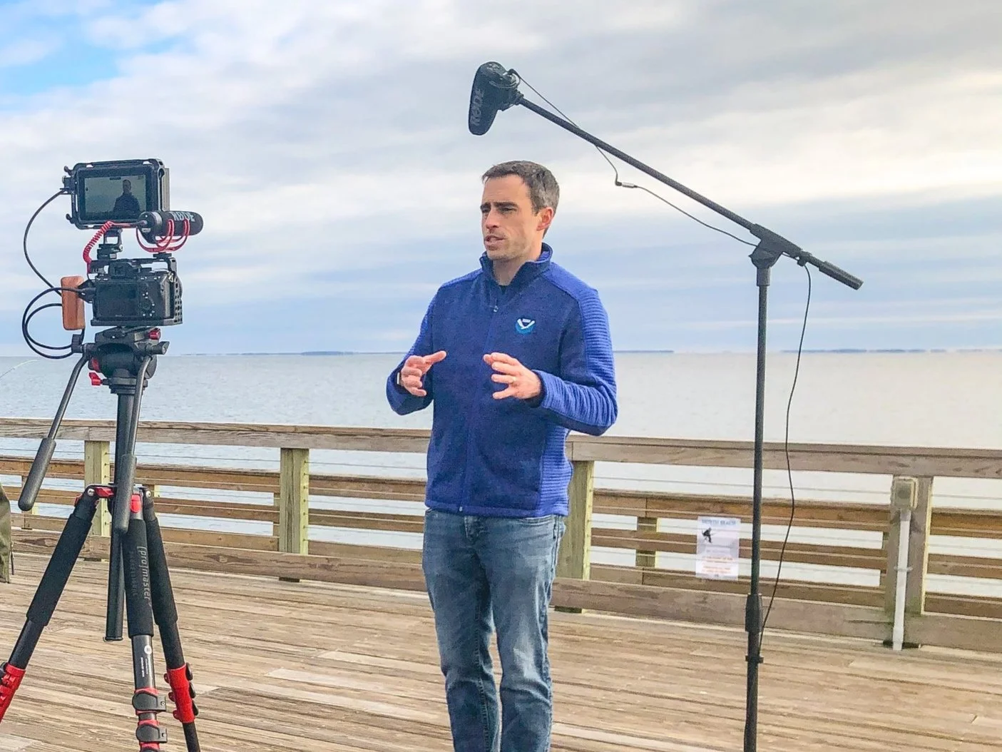 A man in a blue jacket standing on a wooden pier near a body of water, speaking in front of a camera and a boom microphone, with cloudy sky in the background.