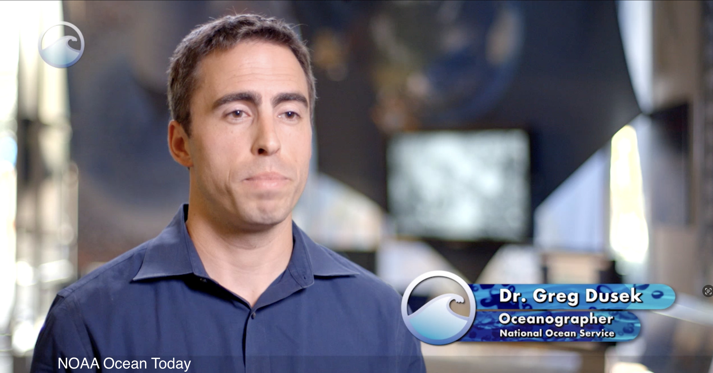 A man in a blue shirt speaking in an interview setting with a graphic overlay identifying him as Dr. Greg Dusek, Oceanographer at the National Ocean Service.