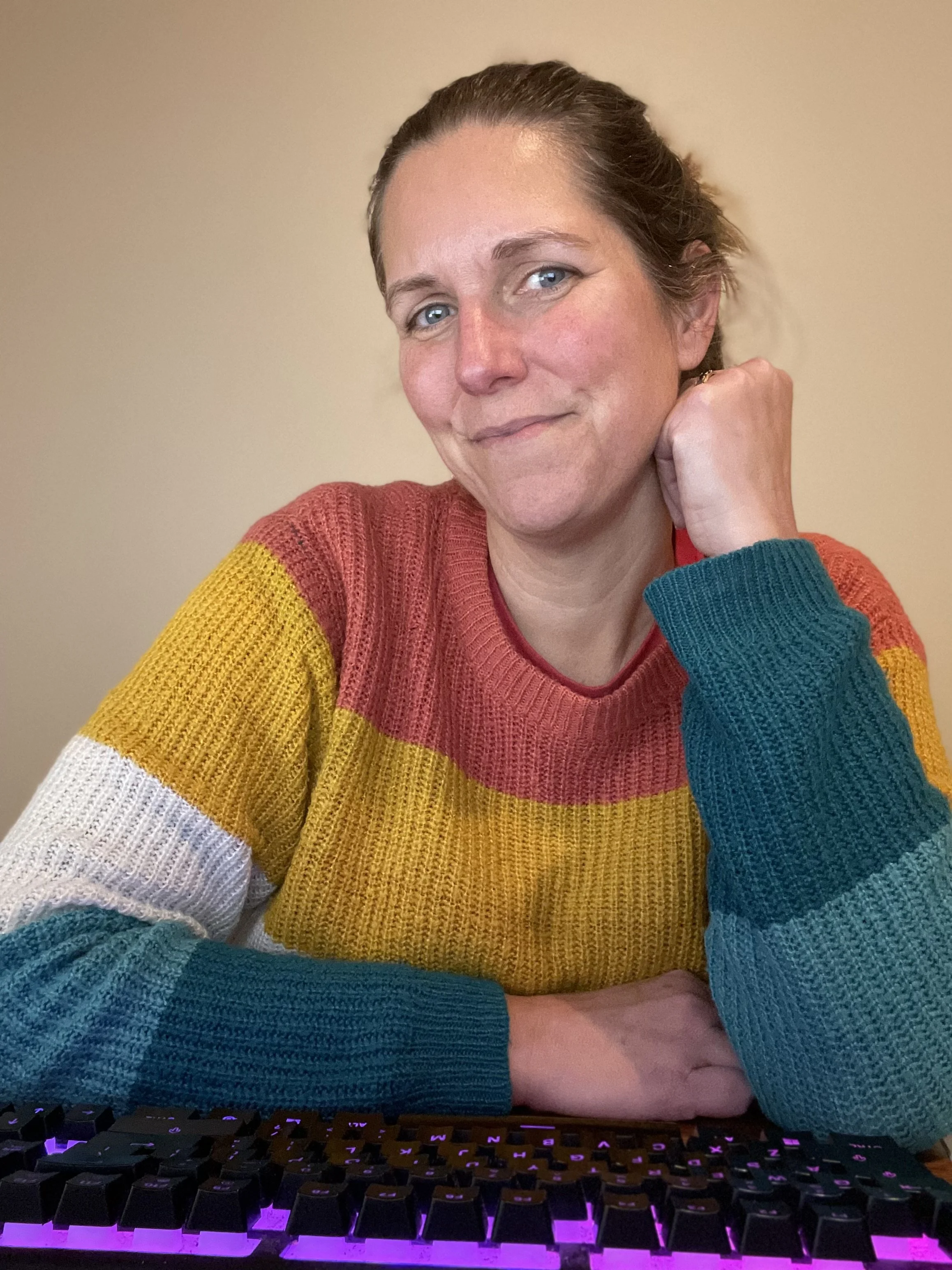 A woman with blue eyes and brown hair tied back, wearing a multicolored sweater with yellow, red, blue, white, and teal sleeves, resting her chin on her hand, sitting in front of a computer keyboard with purple backlit keys.