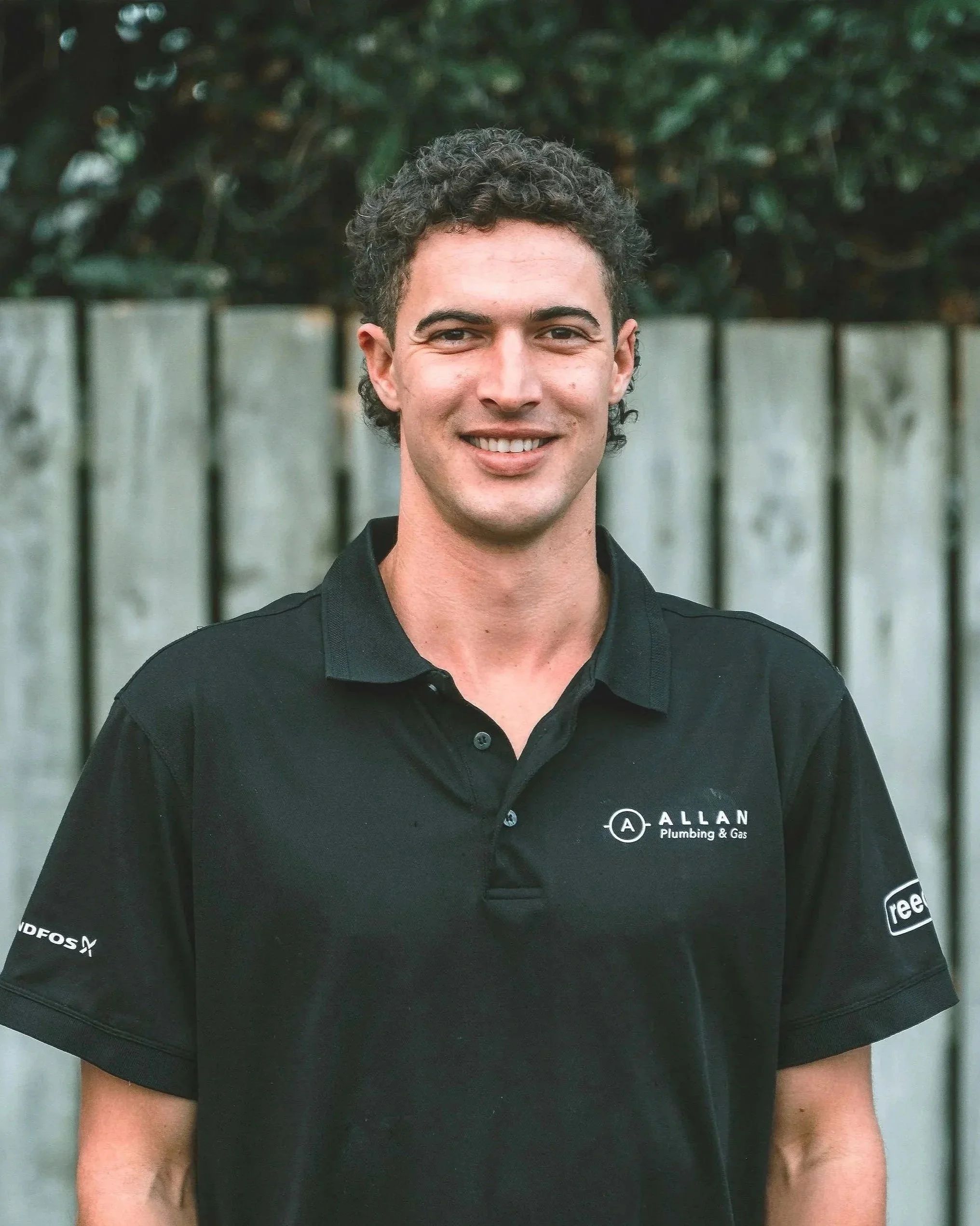 A man with dark brown hair smiling, wearing a black t-shirt that reads 'Lorna Jane Athletic' on the front, standing in front of a wooden fence with some vines.