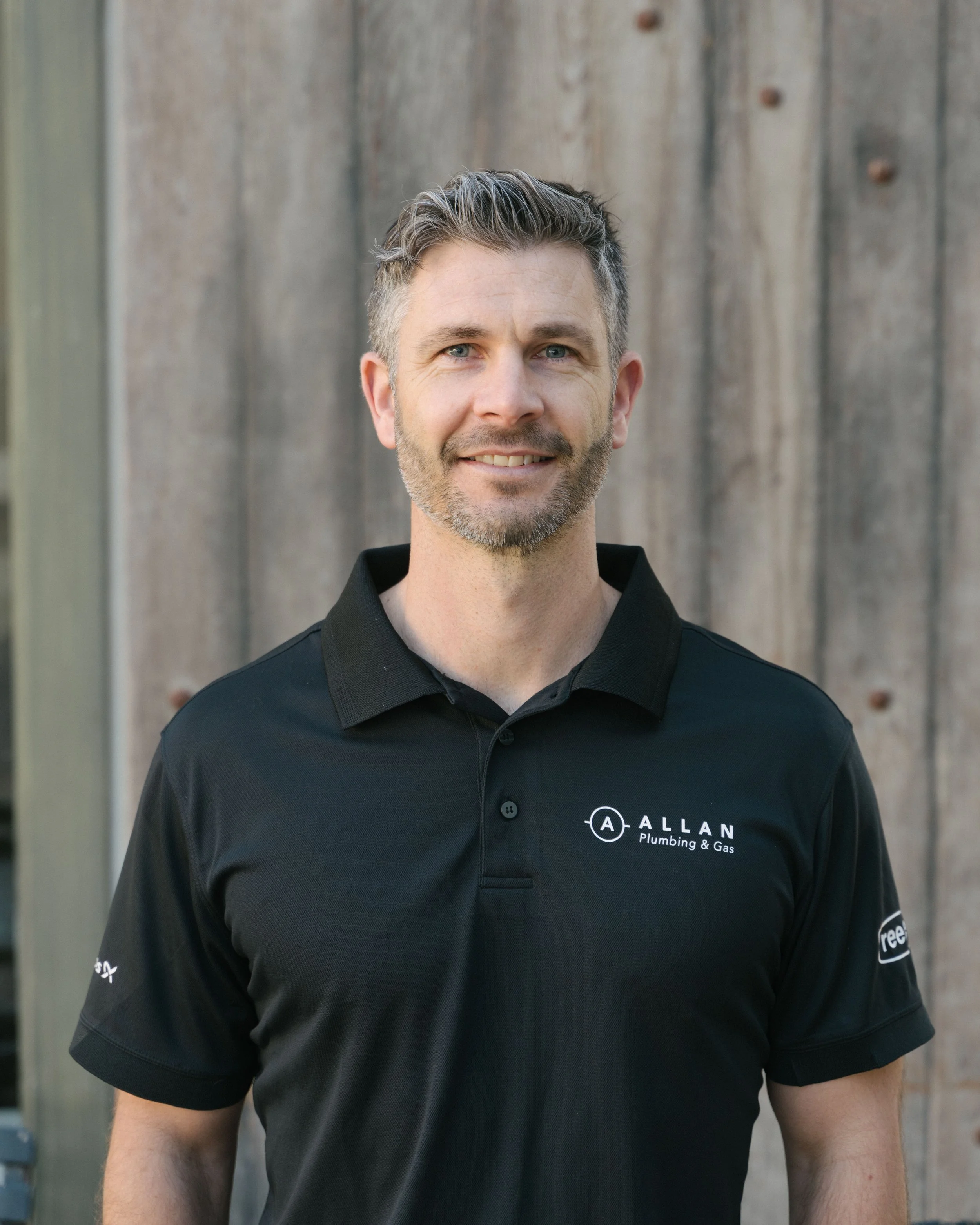 A man with short, dark hair and a beard, smiling, wearing a dark blue polo shirt with 'Allan Plumbing & Gas' logo, standing in front of a wooden wall.
