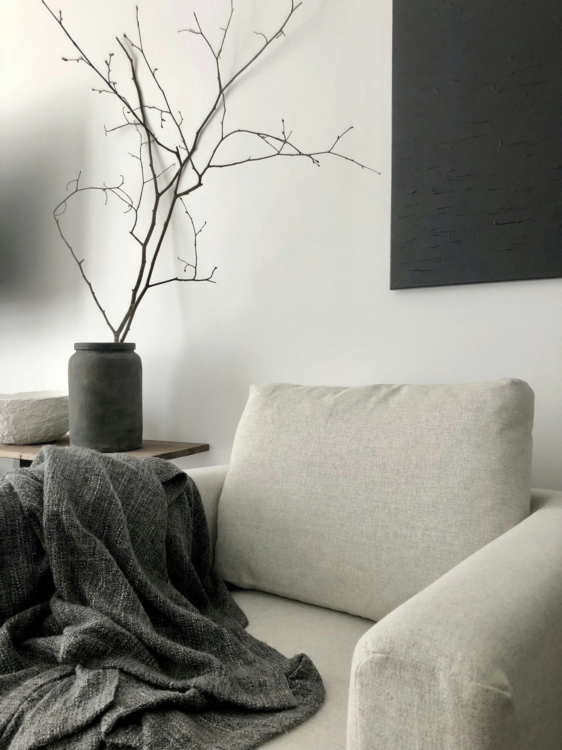 A neutral armchair with a dark gray knit blanket and a vase of bare branches in a quiet modern interior.