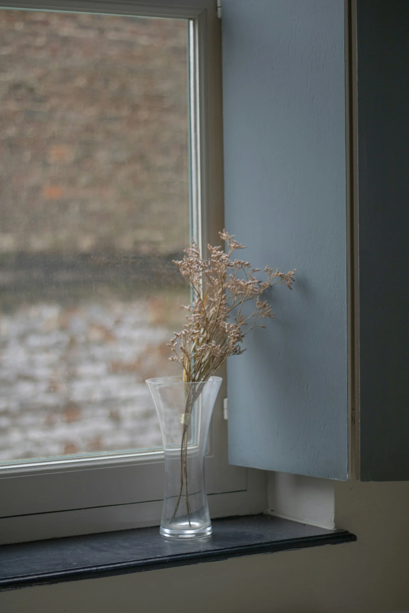 Dried pinkish beige flowers in a clear glass vase placed on a windowsill next to a window with a brick wall outside.