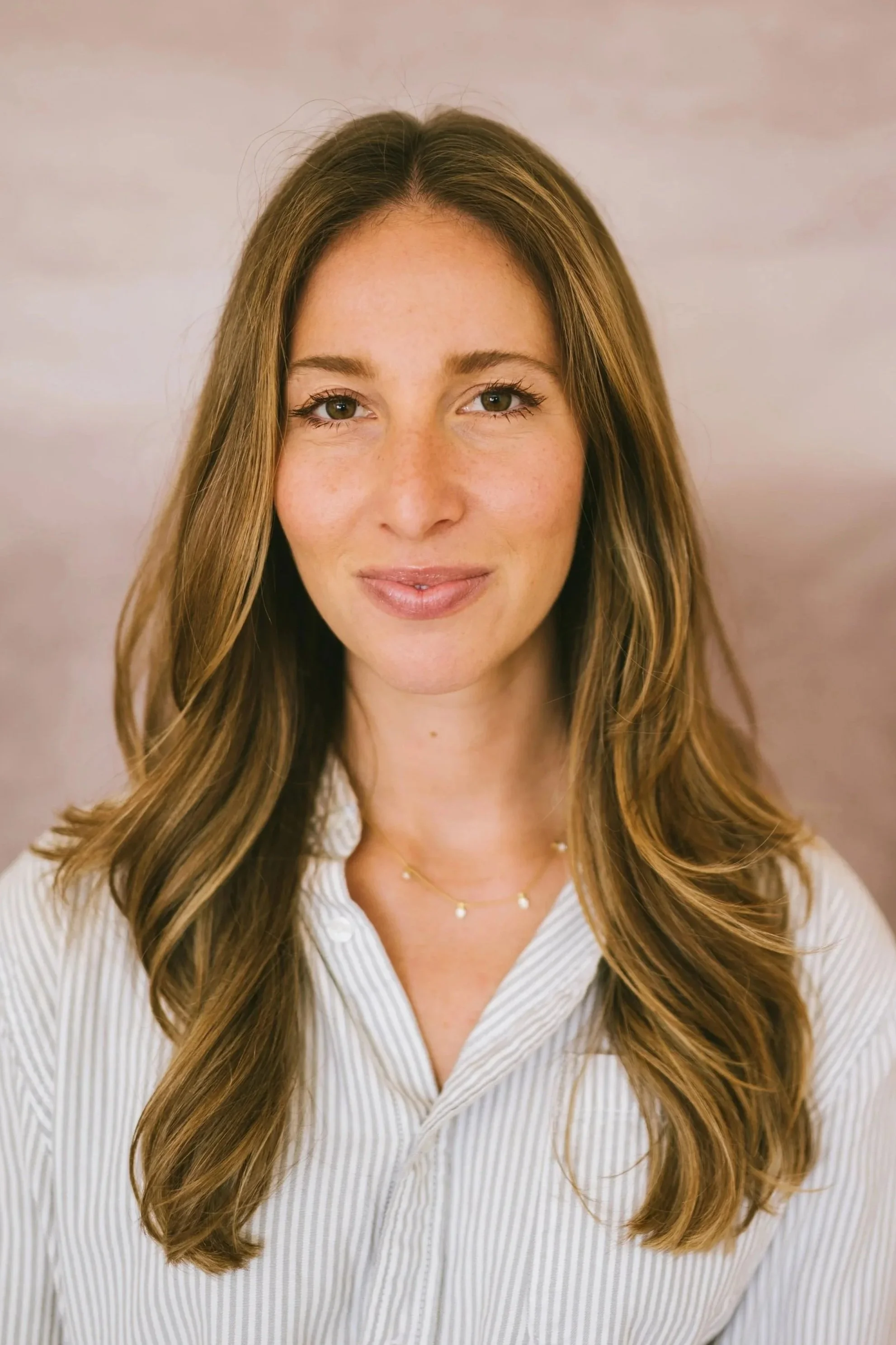 Close-up of a young woman with long, wavy brown hair, wearing a white striped button-up shirt and a delicate gold necklace, smiling softly against a neutral background.