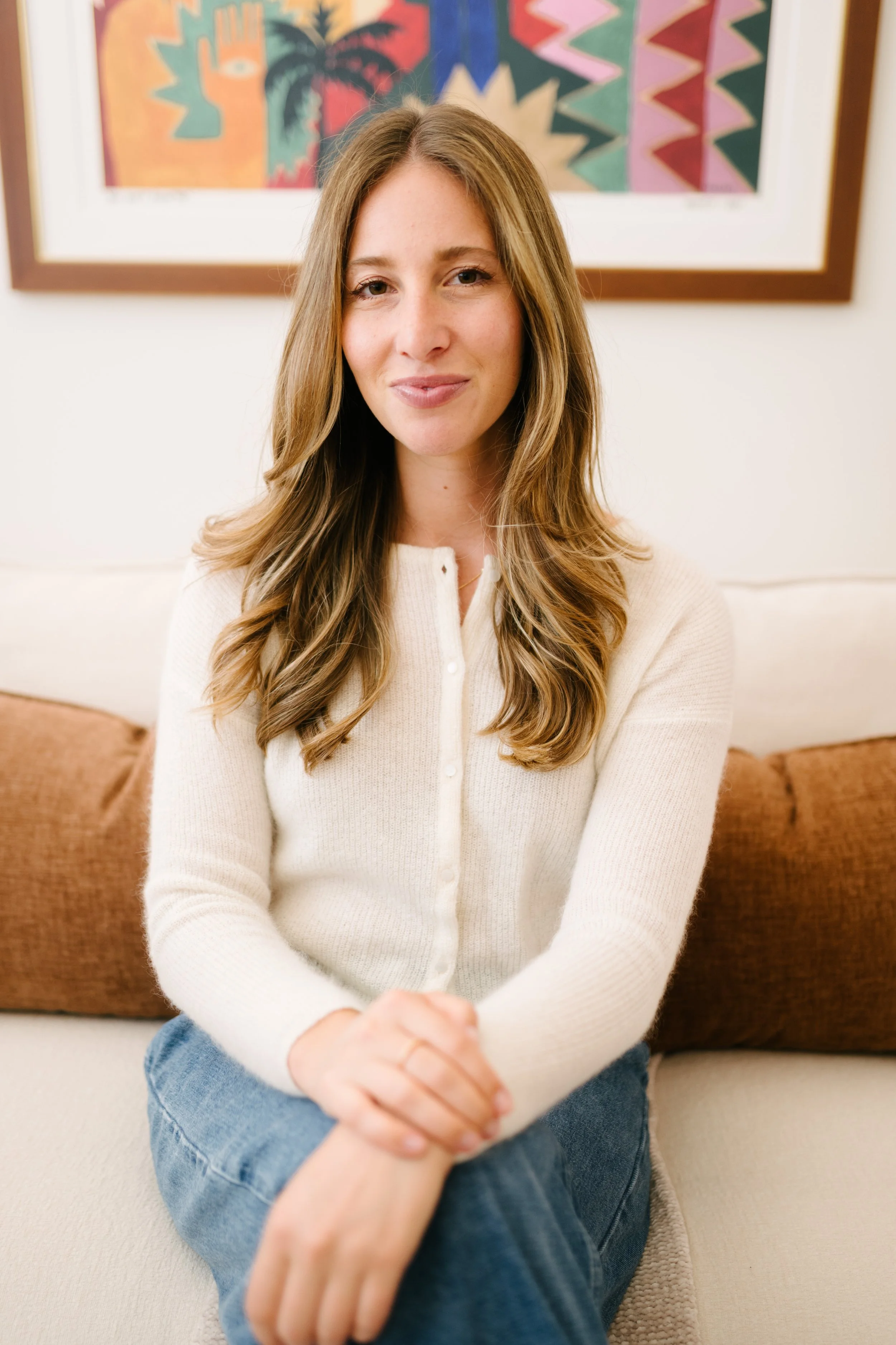 A young woman with long, wavy, light brown hair and fair skin sitting on a beige sofa in front of a colorful abstract painting, wearing a cream-colored cardigan and blue jeans.