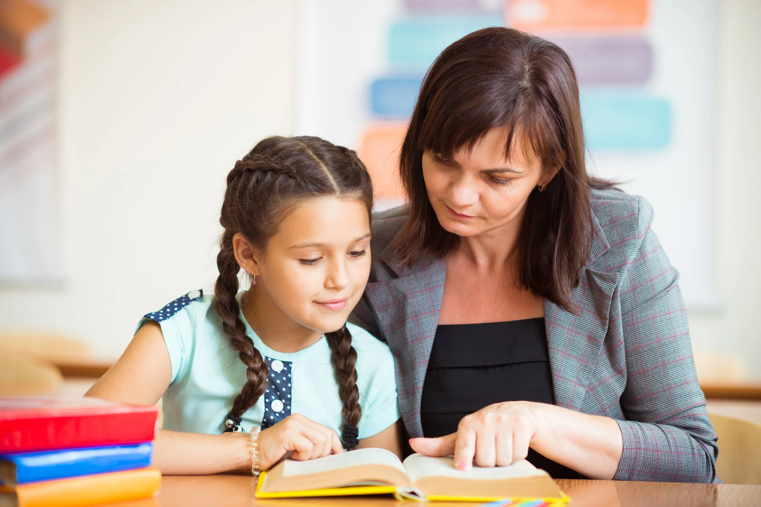 A woman and a young girl sitting at a desk, reading a book together, in a classroom setting with colorful charts on the wall.