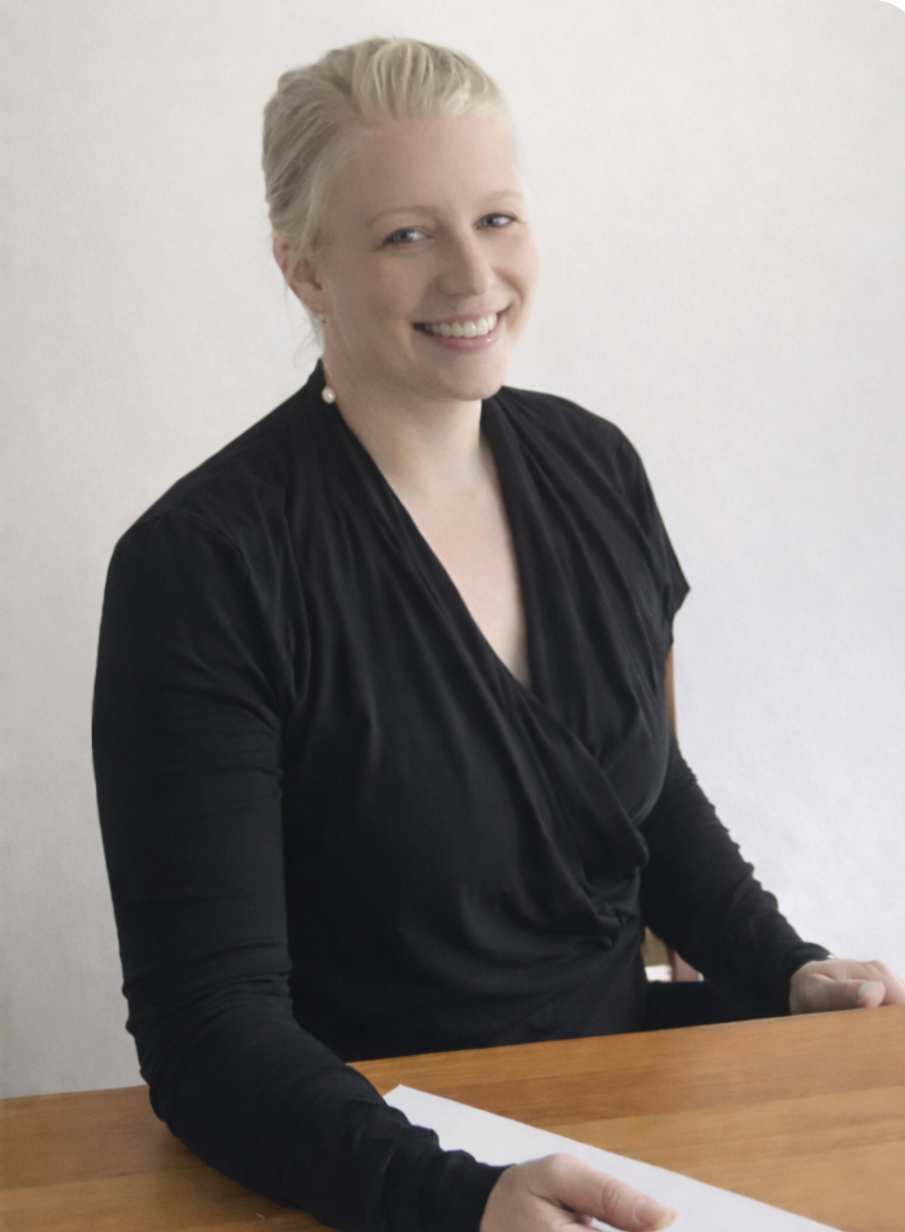A smiling woman with blonde hair tied back, wearing pearl earrings and a black blouse, sitting at a wooden table against a plain light-colored wall.