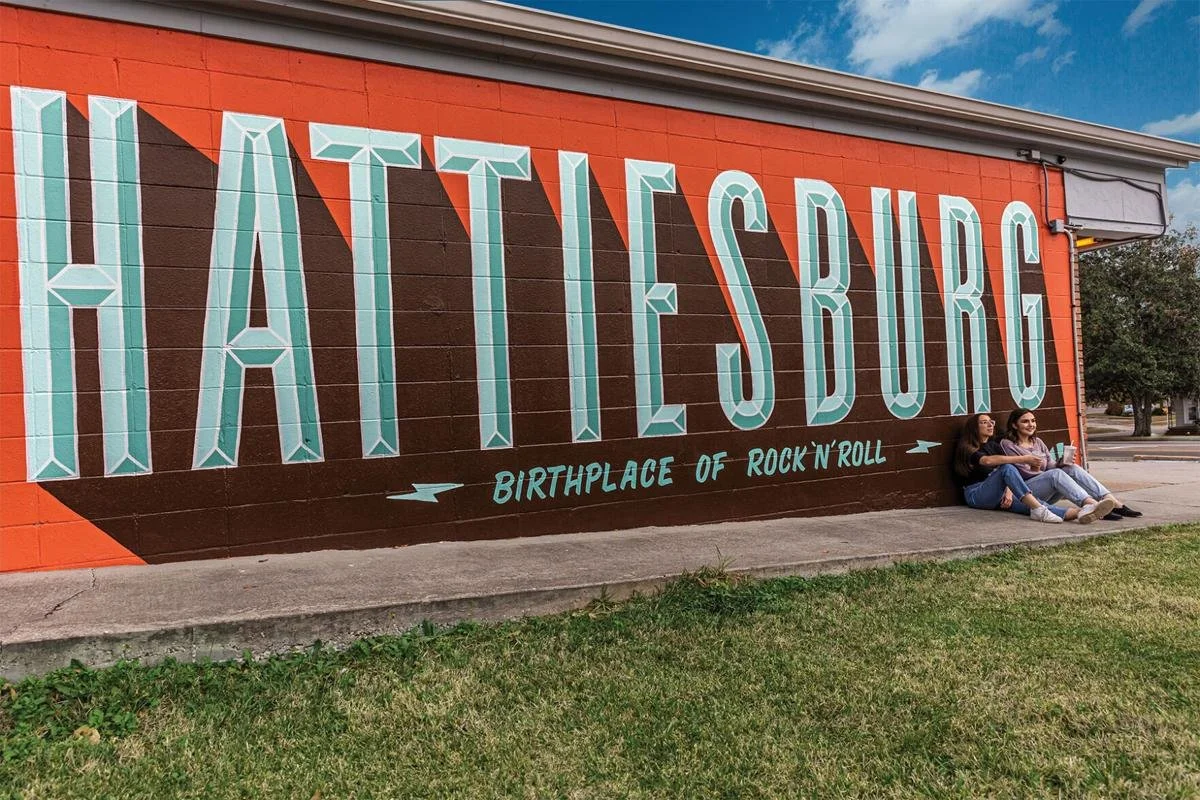 A mural on a brick wall reading 'HATTIESBURG BIRTHPLACE OF ROCK 'N' ROLL' with two women sitting on the sidewalk in front of it.