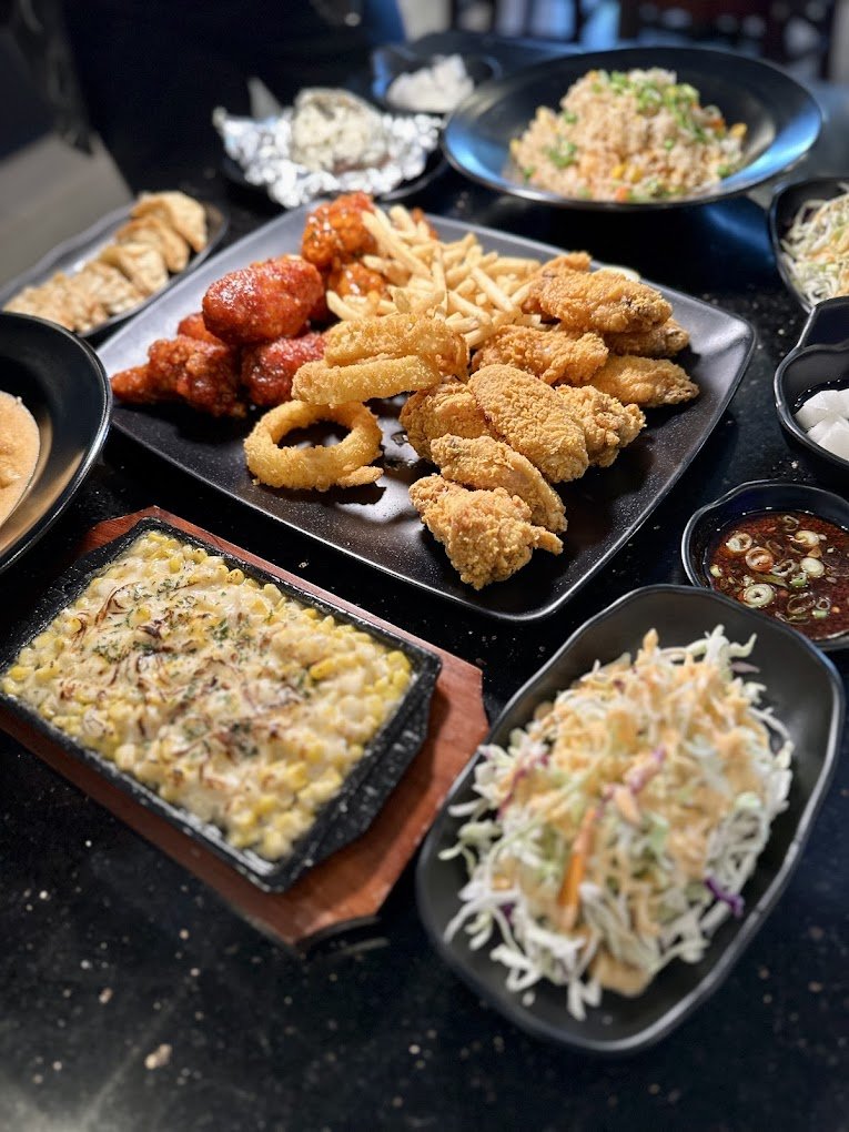 A variety of dishes on a table including fried chicken, onion rings, fries, buffalo wings, coleslaw, fried rice, cheesy corn, and dumplings on black plates.