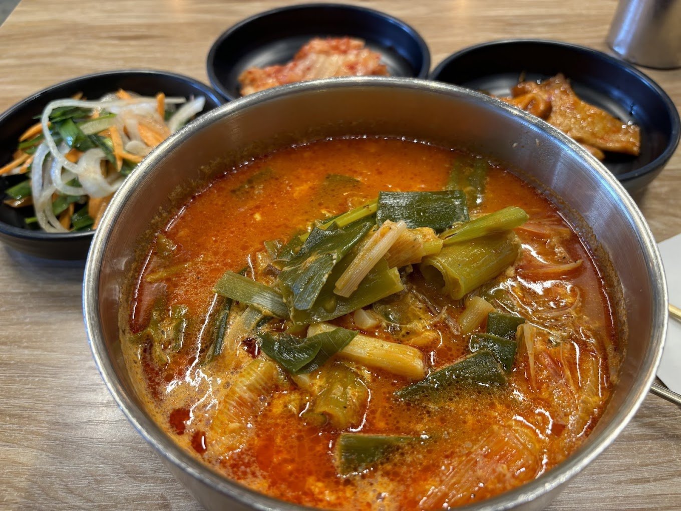 A bowl of Korean kimchi stew with vegetables, accompanied by side dishes of kimchi, seasoned vegetables, and fried fish cake on a wooden table.