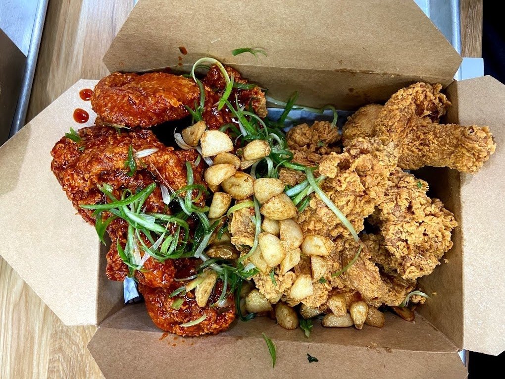 A takeout box containing Korean fried chicken. The box includes crispy fried chicken on one side and spicy glazed fried chicken on the other, garnished with green onions and roasted garlic.