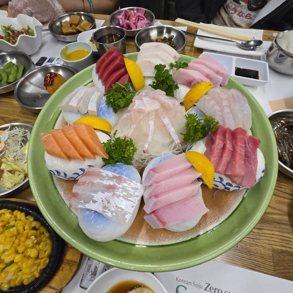 A colorful sashimi platter with various types of sliced raw fish, garnished with parsley and lemon wedges, served on a green plate. Accompanied by side dishes and sauces on a wooden table.
