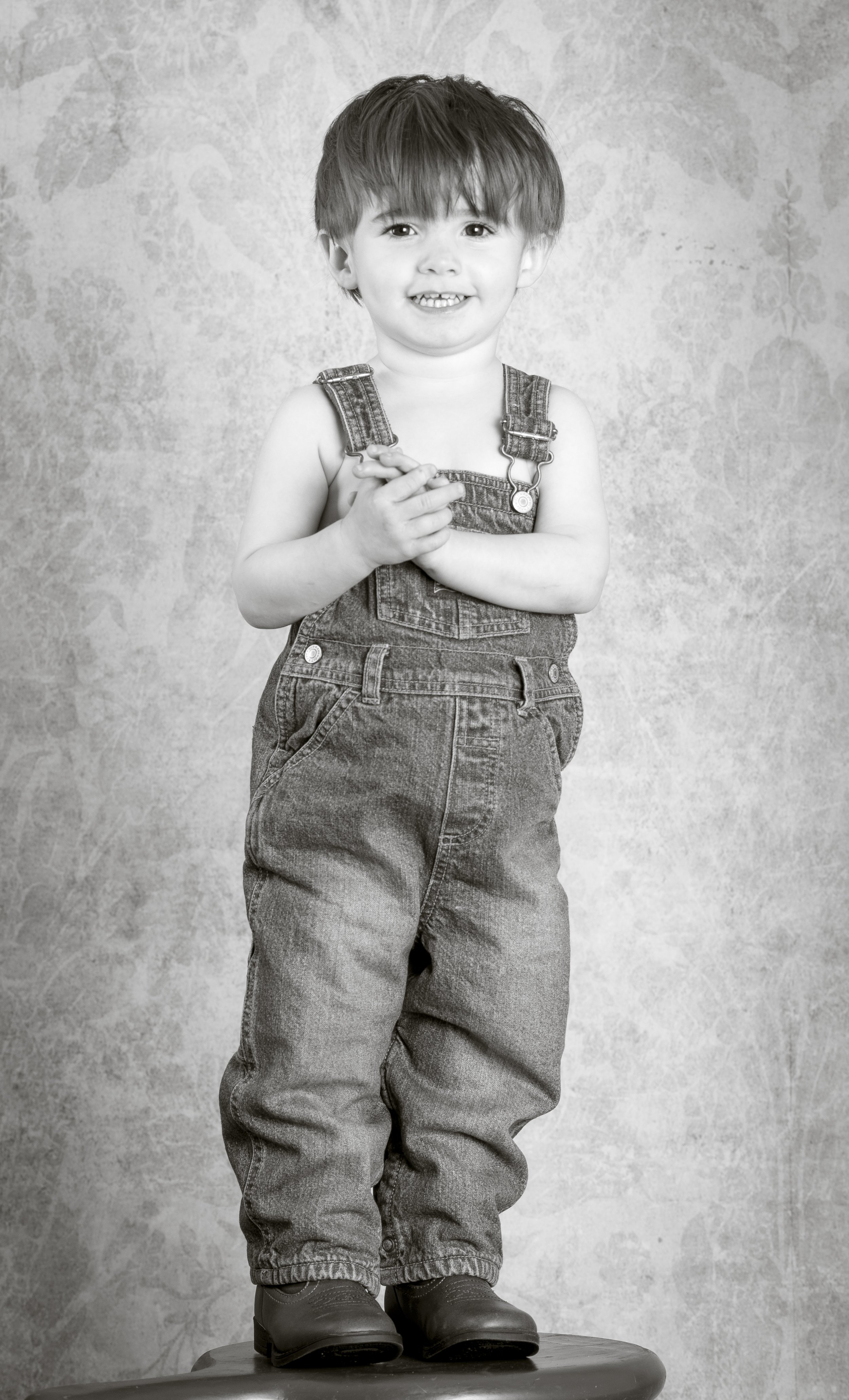 A young boy with messy hair, wearing overalls and boots, standing on a small platform and smiling at the camera.