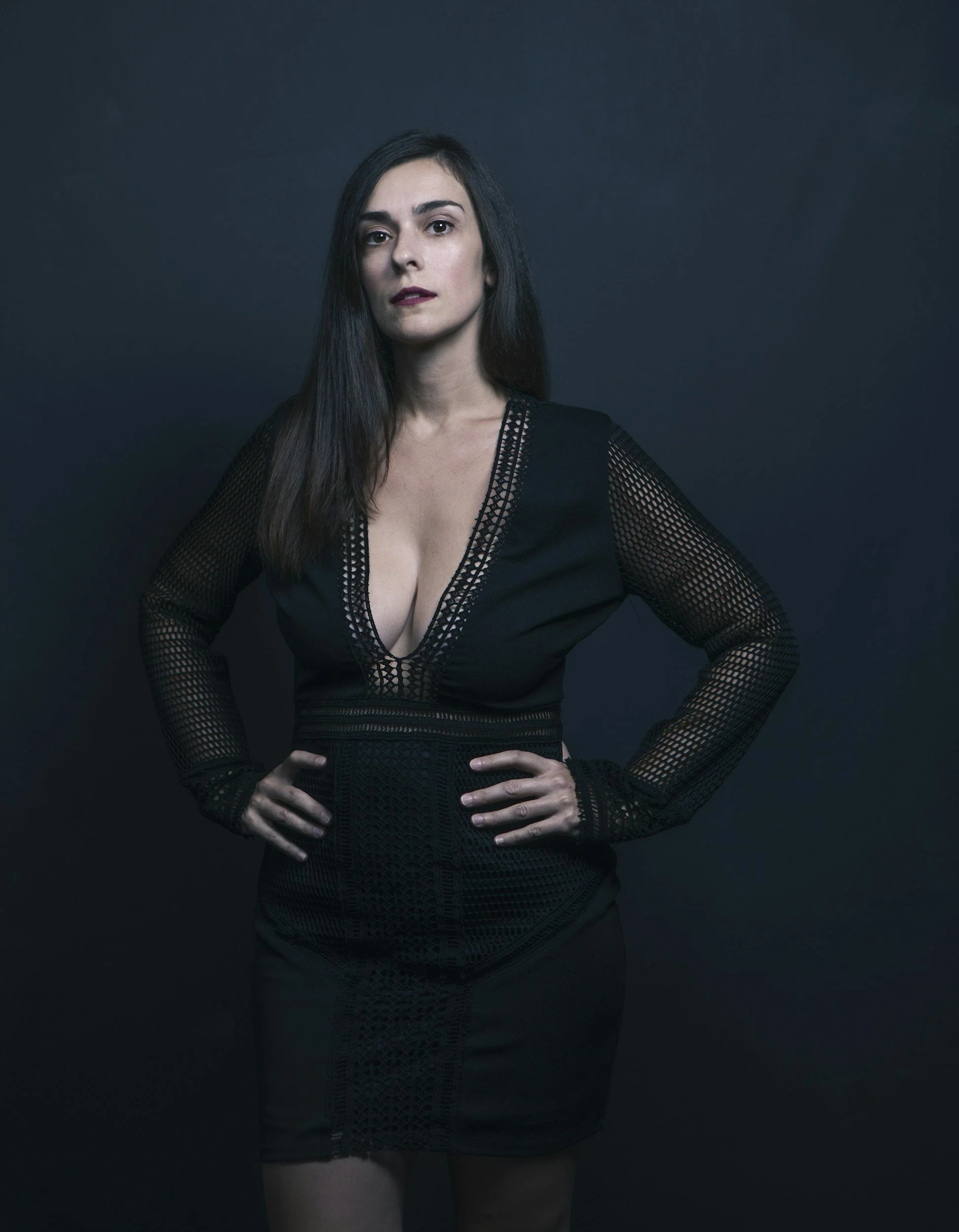 A woman with long dark hair and fair skin wearing a black dress with lace and mesh details, standing against a dark background with hands on her hips.