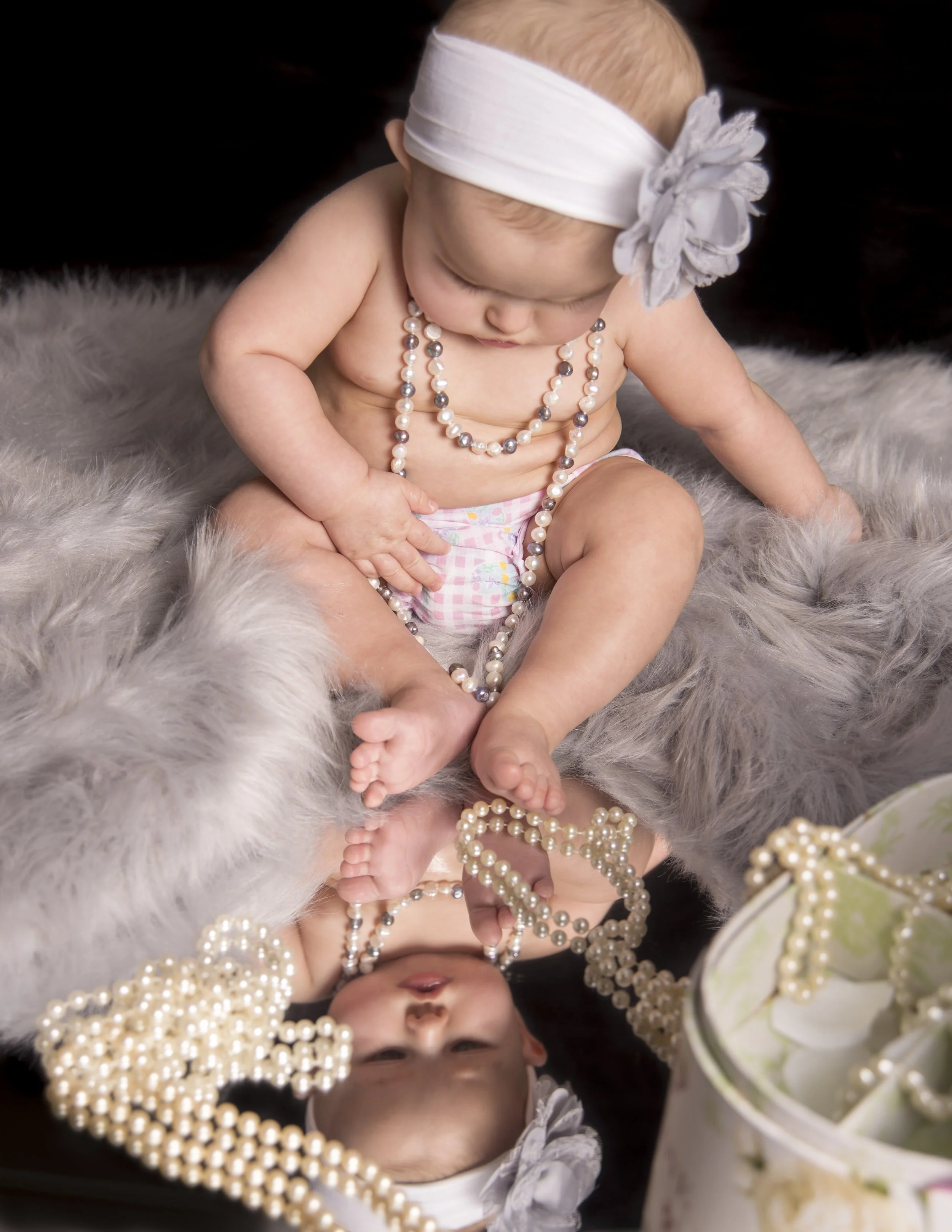 A baby girl sitting on a fluffy gray blanket, wearing a white headband with a flower, beads, and a diaper, with a reflection of her in a shiny surface below.
