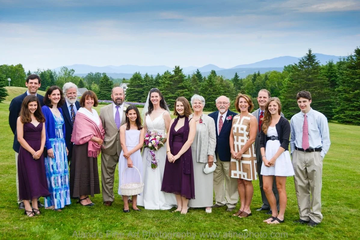 Group of people, including women and men of various ages, dressed in formal and semi-formal attire, standing outdoors on a grassy field with trees and mountains in the background, during daytime.