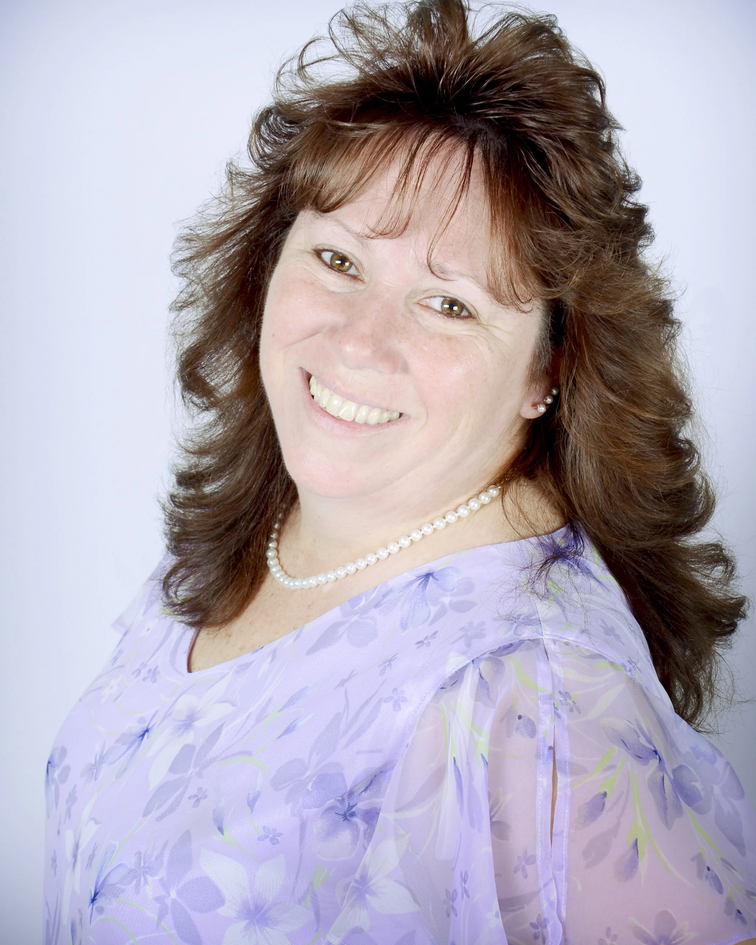 A woman with shoulder-length brown hair, smiling, wearing a pearl necklace and a light purple floral blouse against a plain white background.