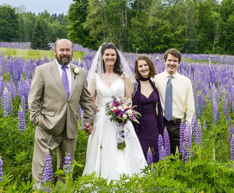 A bride holding a bouquet stands with three people, likely her family, in a field of purple flowers with green trees in the background.
