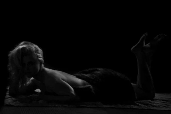 A person lying on their stomach on a surface with a dark background, partly illuminated, with one arm supporting their head and their legs bent at the knees.