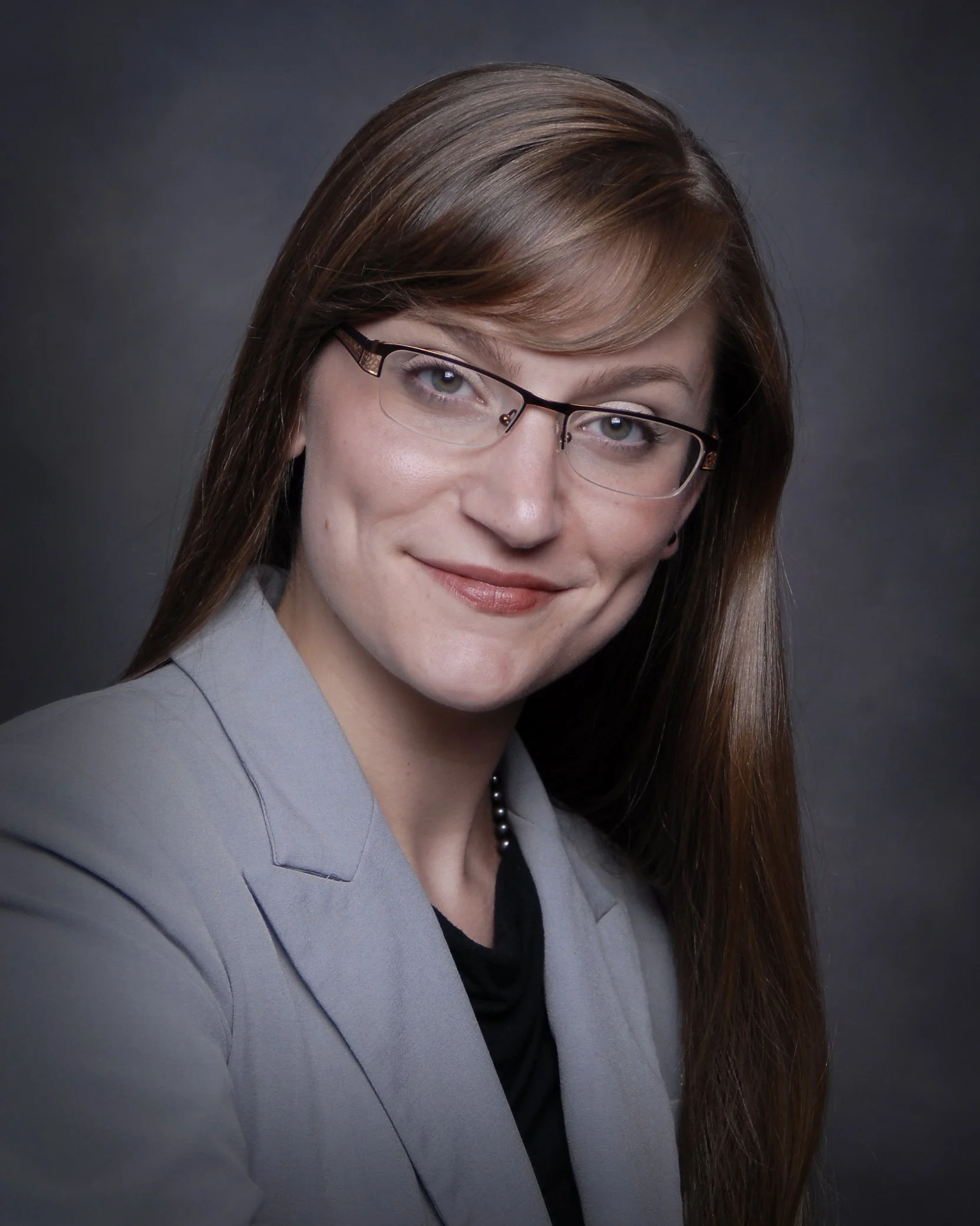 A professional headshot of a woman with long brown hair, wearing glasses, a gray blazer, and a black top, smiling softly against a dark background.