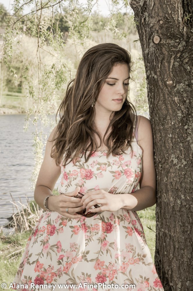 A young woman in a floral dress stands next to a tree near a body of water, holding sunglasses.