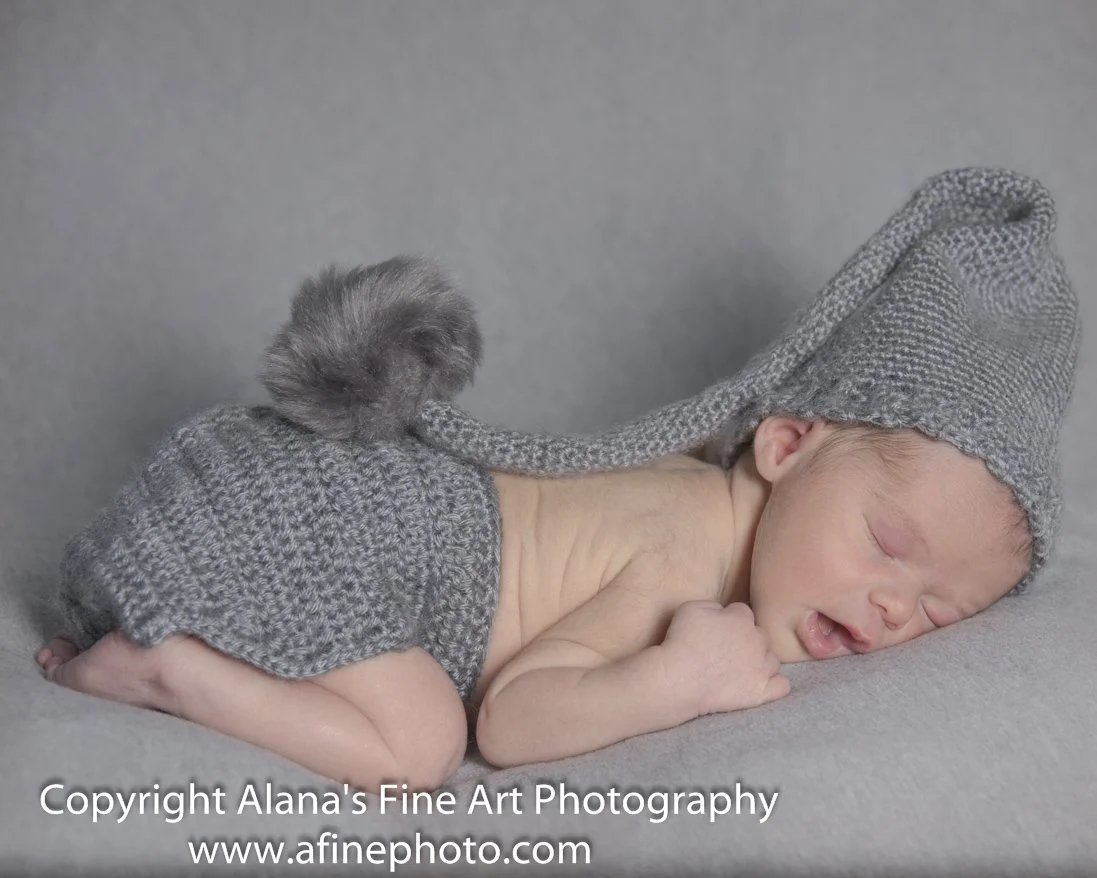 A sleeping baby dressed in a gray knit hat and matching gray knit diaper cover with a pom-pom, lying on its side on a gray surface.