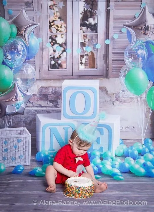 Child wearing a party hat and red shirt sitting on the floor with a birthday cake in front of him, surrounded by blue and green balloons and star-shaped balloons, with large blocks spelling "ONE" in the background, and a decorated wall with a window.