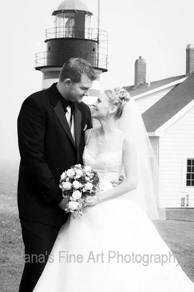 A bride and groom standing closely outdoors near a lighthouse and a white building, smiling at each other after their wedding.