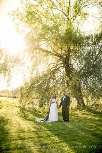A bride in a white wedding dress and groom in a suit standing beneath a large tree in a grassy field during sunset.