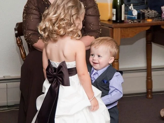 A young girl in a white dress with a large black bow interacting with a smiling young boy in a suit, with an adult woman behind them, in a room with a wooden table and drinks.
