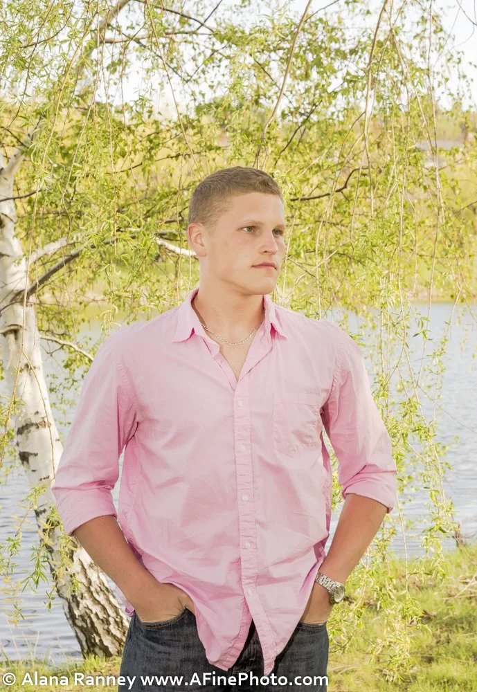A young man with light skin and short hair wearing a pink button-up shirt, dark jeans, a watch, and a necklace, standing outdoors near a body of water with a tree and green foliage in the background.