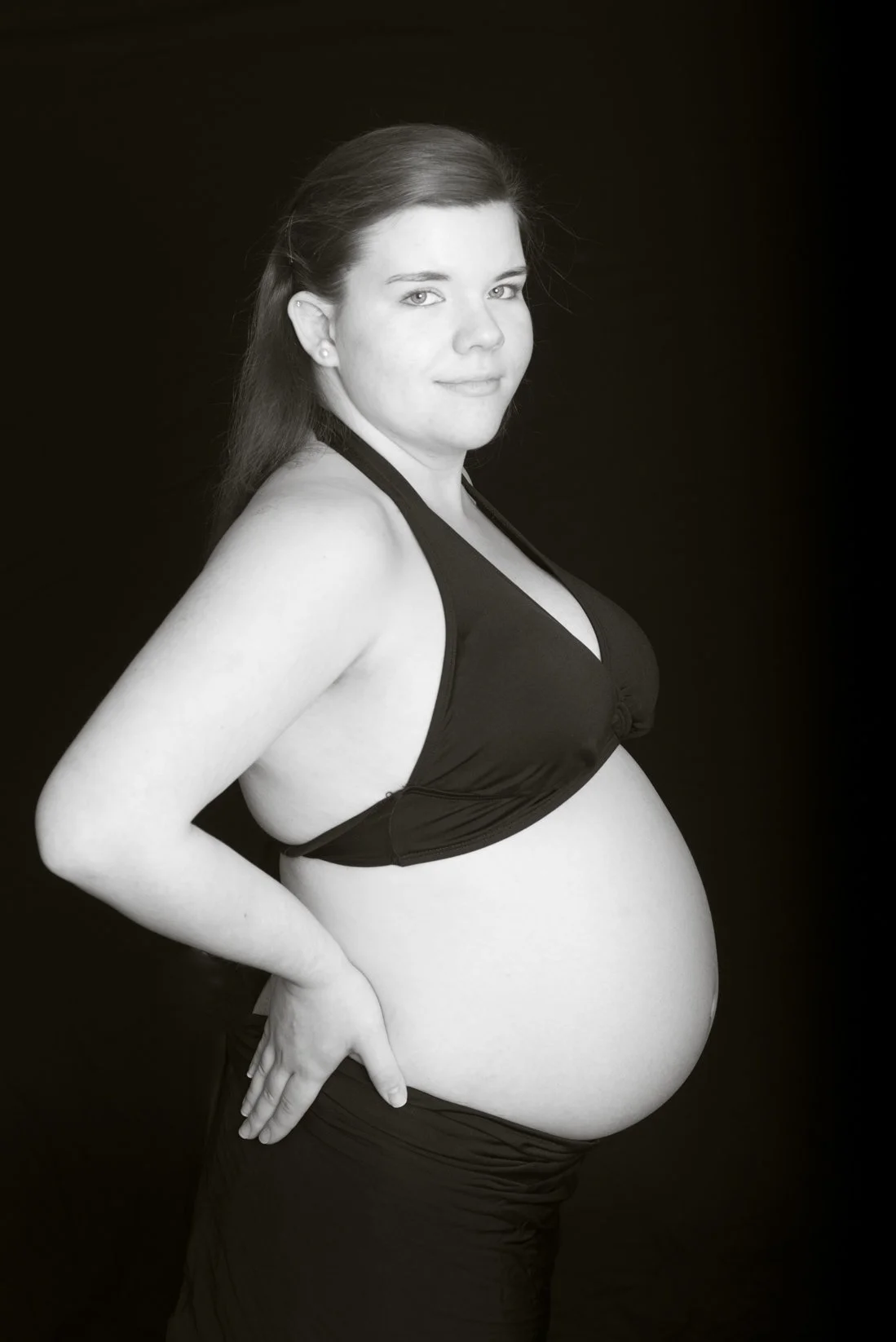 Black and white photo of a pregnant woman in a sleeveless top, standing against a dark background, looking towards the camera with a slight smile.