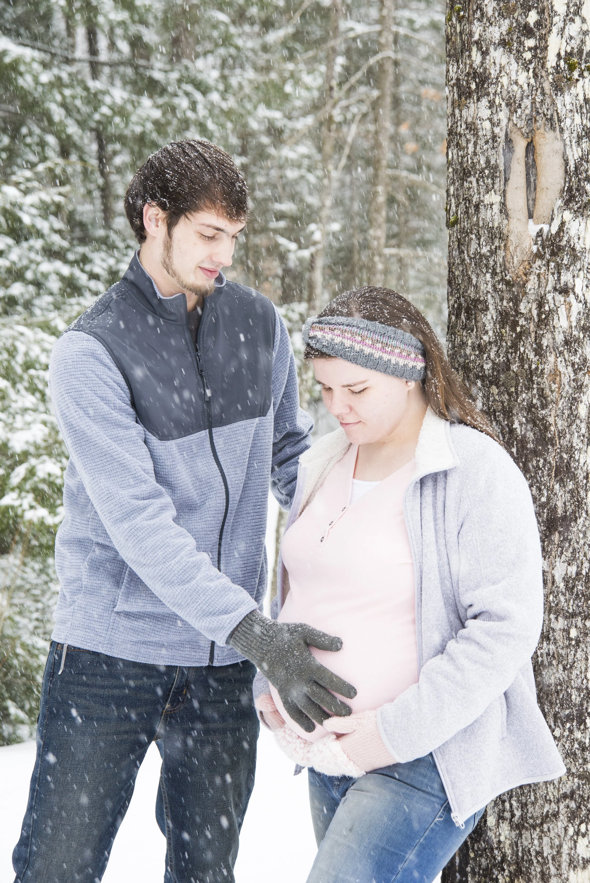 A man and a pregnant woman are standing outdoors in a snowy forest. The man is gently touching the woman's pregnant belly, and both are looking down, with the woman leaning against a tree.