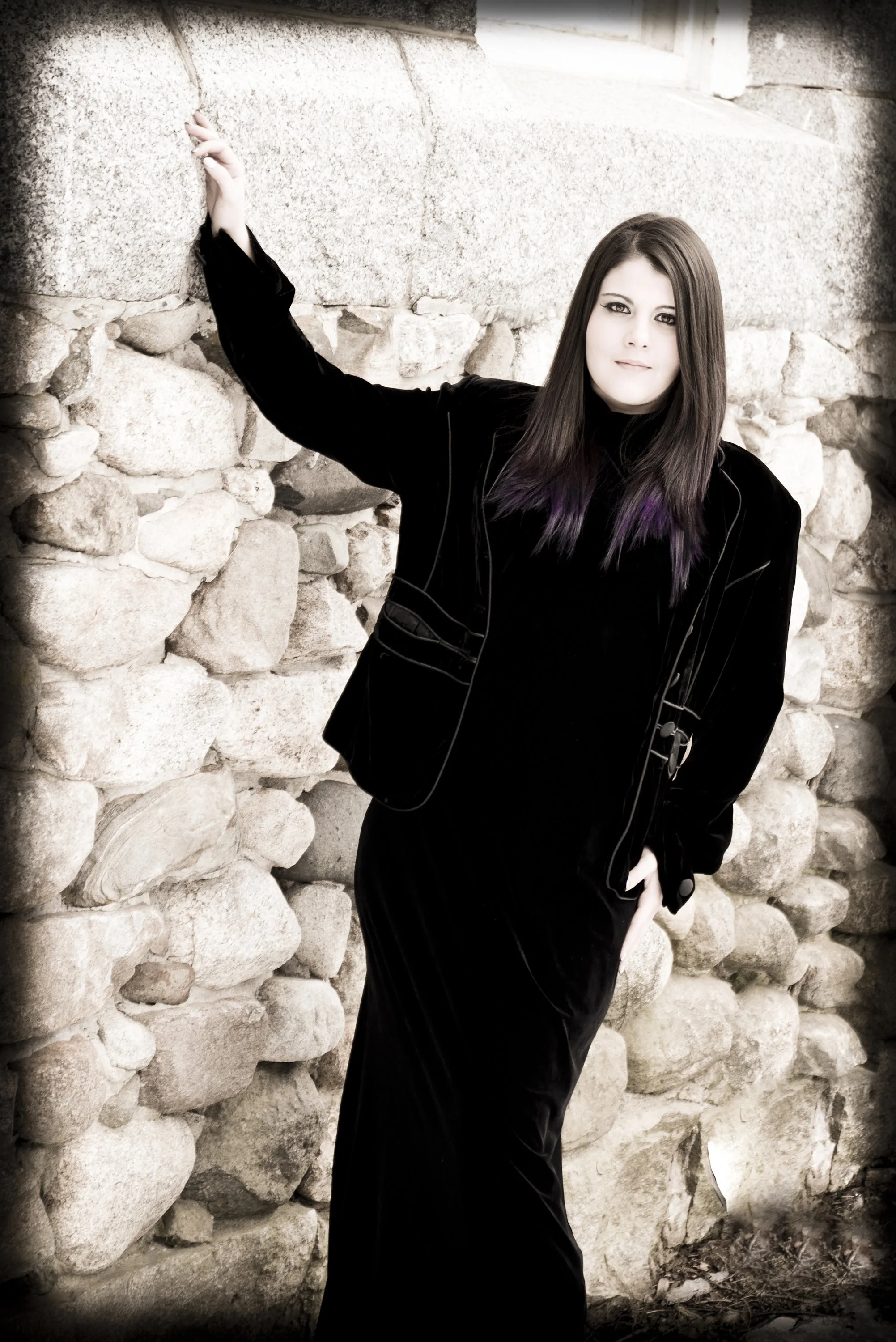 A woman with straight dark hair and purple tips, wearing a black outfit, stands against a stone wall, looking at the camera.