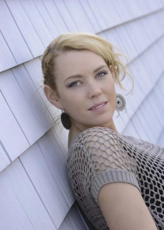 A woman with blonde hair and blue eyes leaning against a white wooden wall, wearing a crochet top and large earrings.