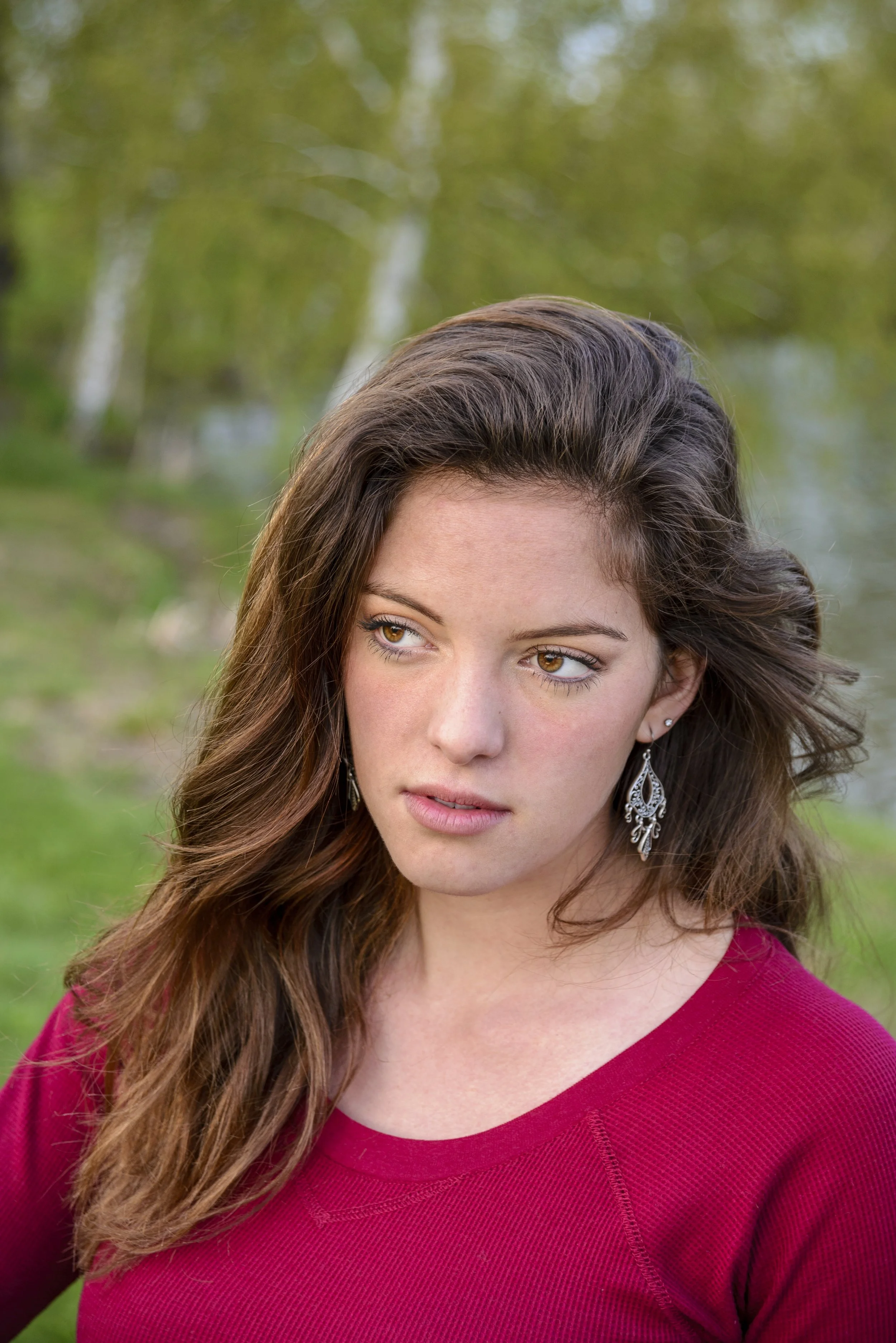 A woman with long, wavy brown hair and brown eyes outdoors by a river, wearing a red top and decorative earrings.